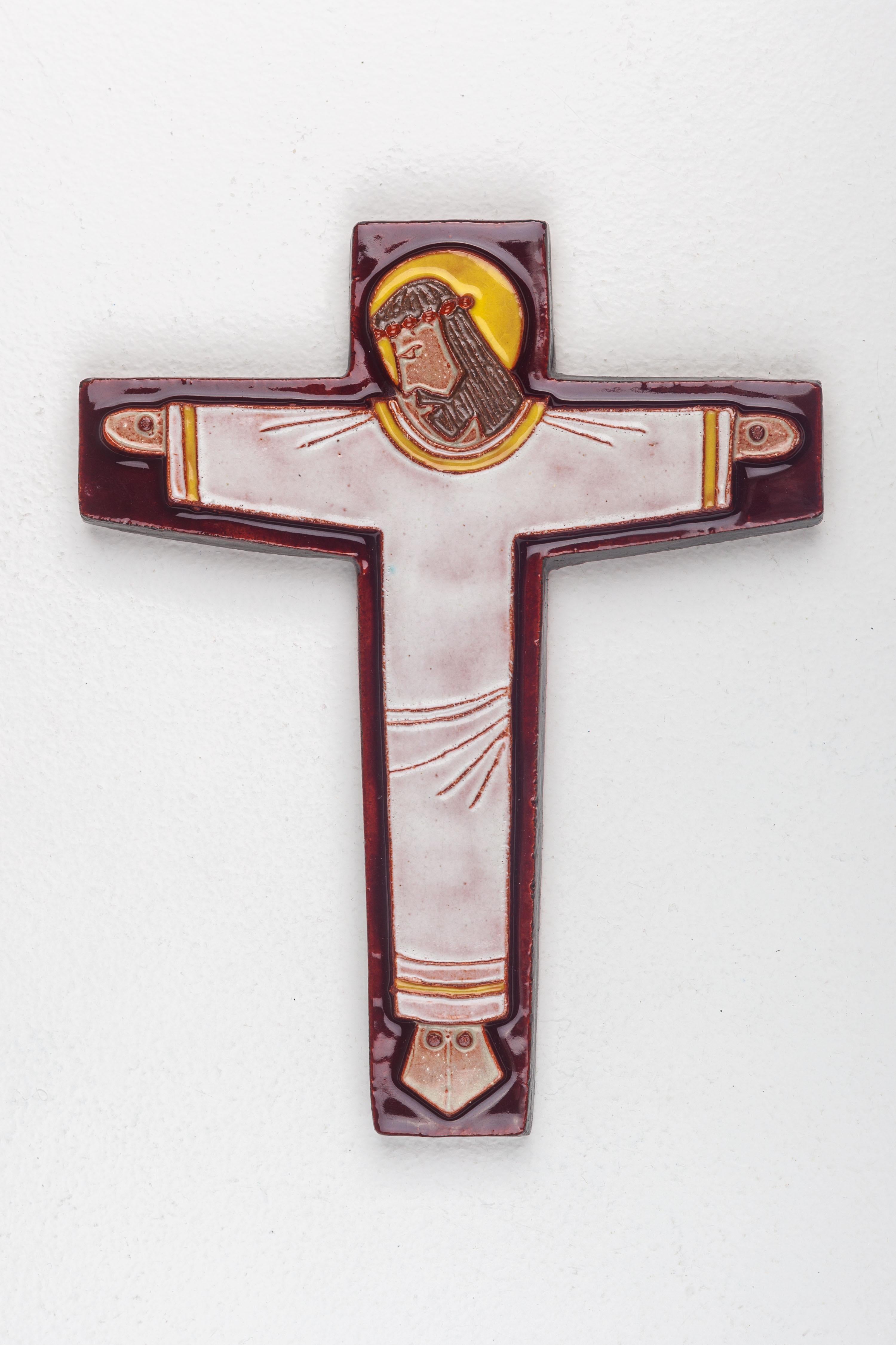 A midcentury European ceramic cross with an archetypal Christ figure adorned by a yellow halo. An interesting combo of color and texture with a variety of earth tones and a high gloss glaze. 

This cross mirrors the architectural landscapes of the