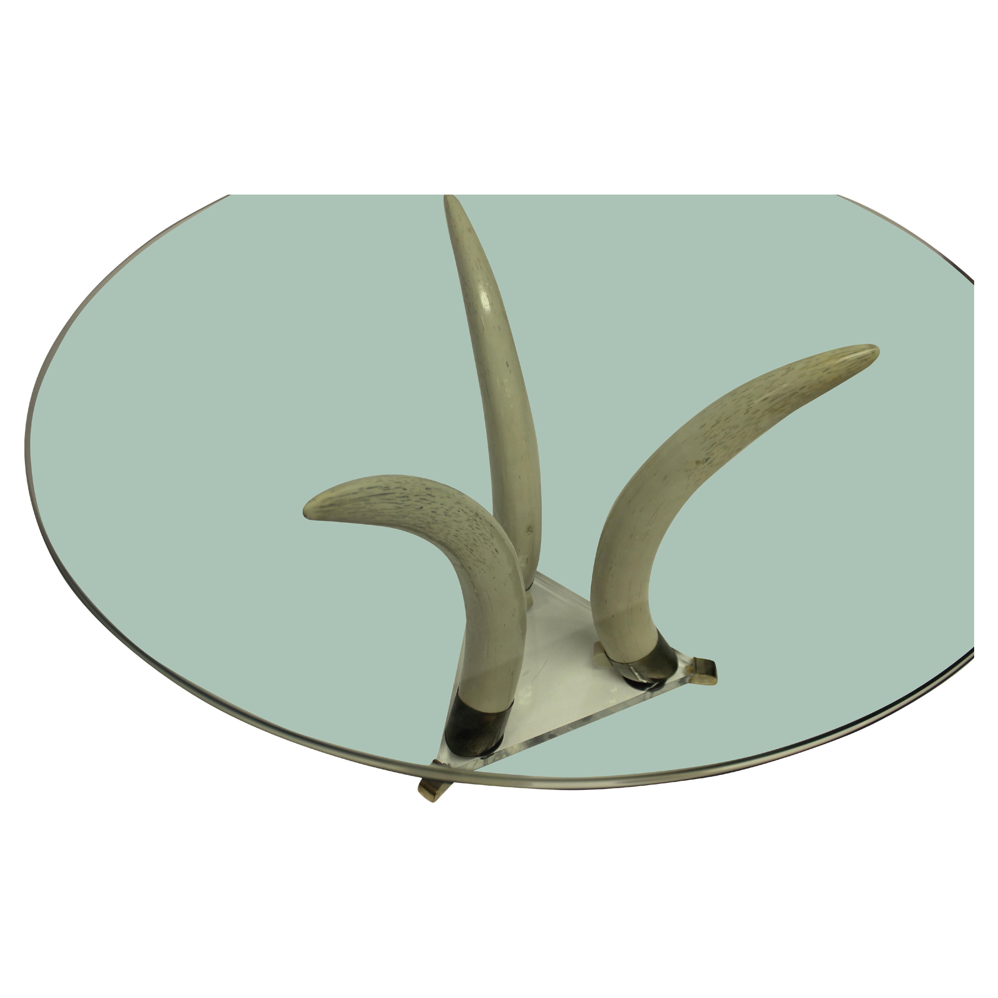 A French mid-century faux tusk centre table. On an acrylic triangular base with brass feet, the three life like tusks, supporting a circular clear plate glass top.