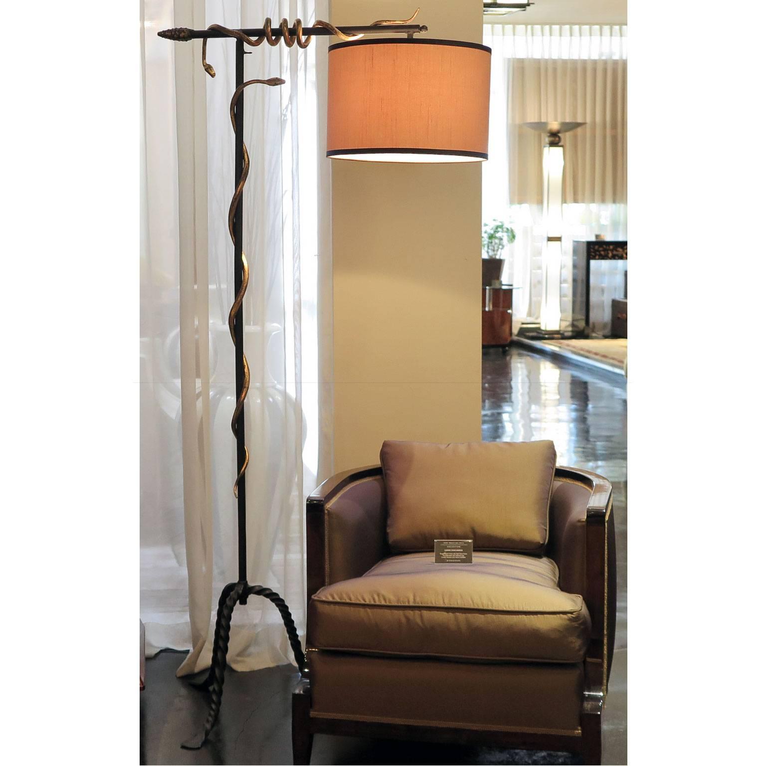 Unusual floor lamp with a black metal stem and three twisted black wrought iron legs. Two gilded snakes circle around the stems and a gilded apron detail decorates the end. A round silk shade hangs from the top stem. This gilded snake floor lamp has