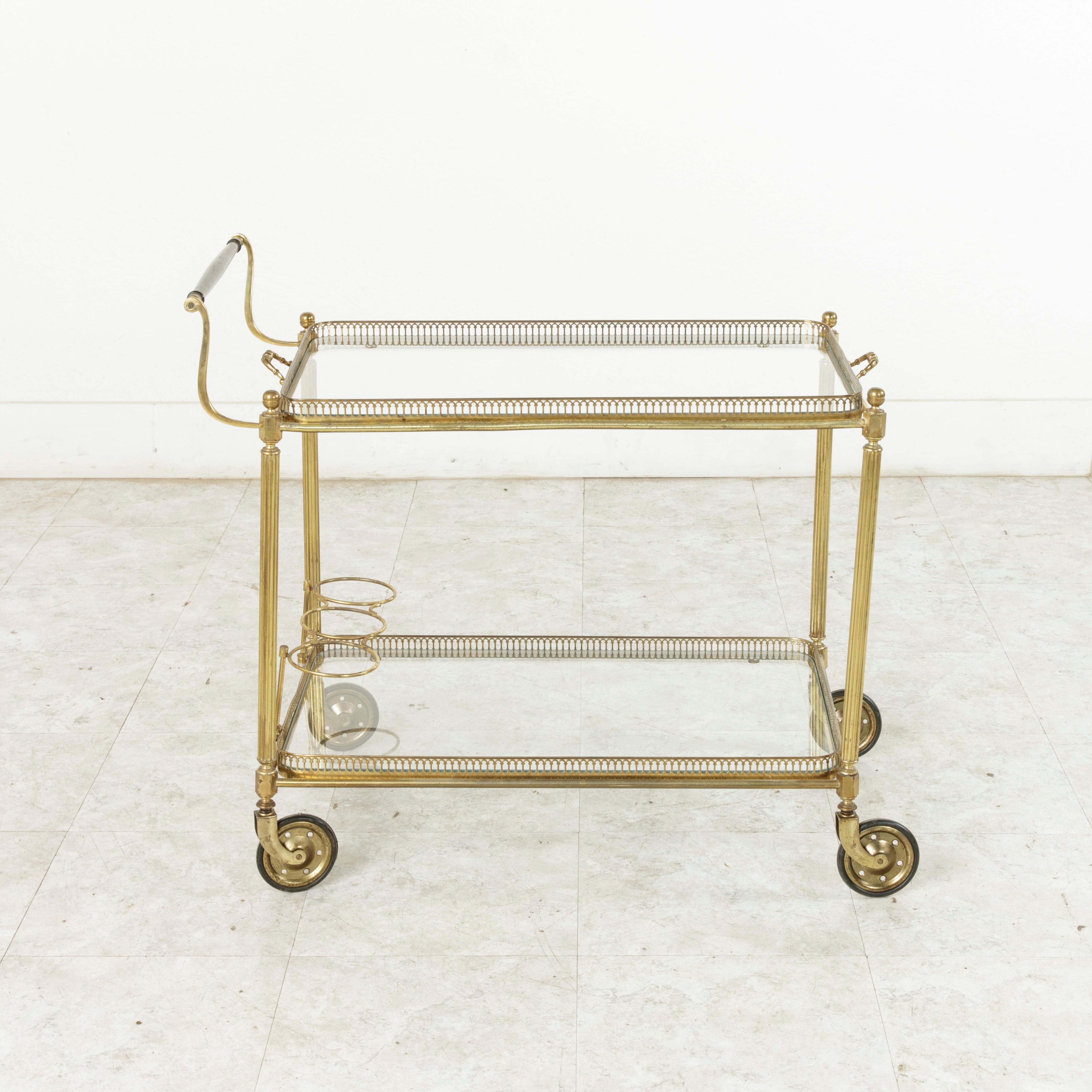 This midcentury French Louis XVI style brass bar cart features a mahogany wood handle and two glass shelves surrounded by pierced brass galleries. The upper shelf is removable and doubles as a serving tray with a handle on each side. The lower shelf