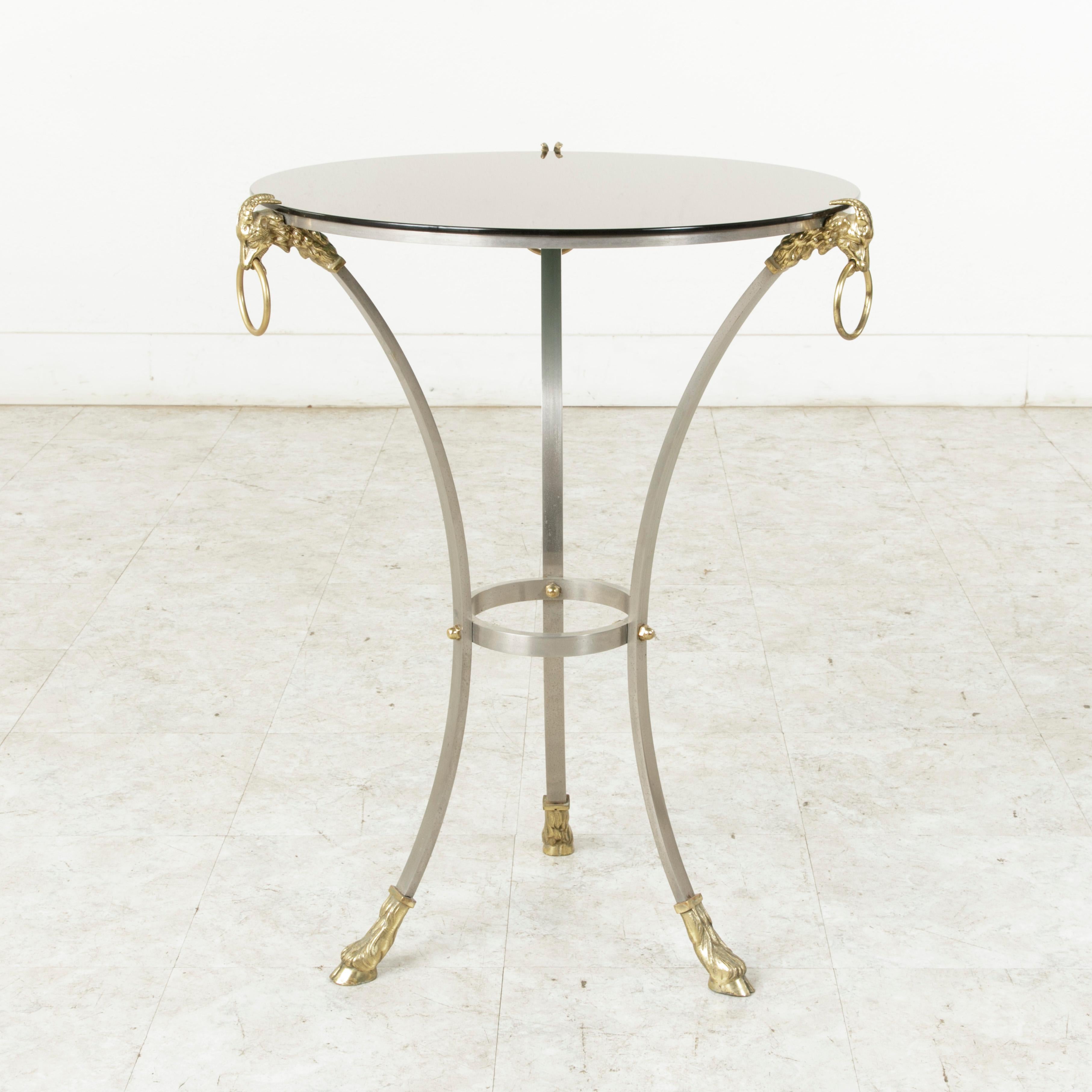 This midcentury French steel gueridon or side table is attributed to the design house Maison Charles. It features a tripod base formed by three legs capped with brass rams' heads bearing brass rings and finished with brass hoof feet. A bronze mirror