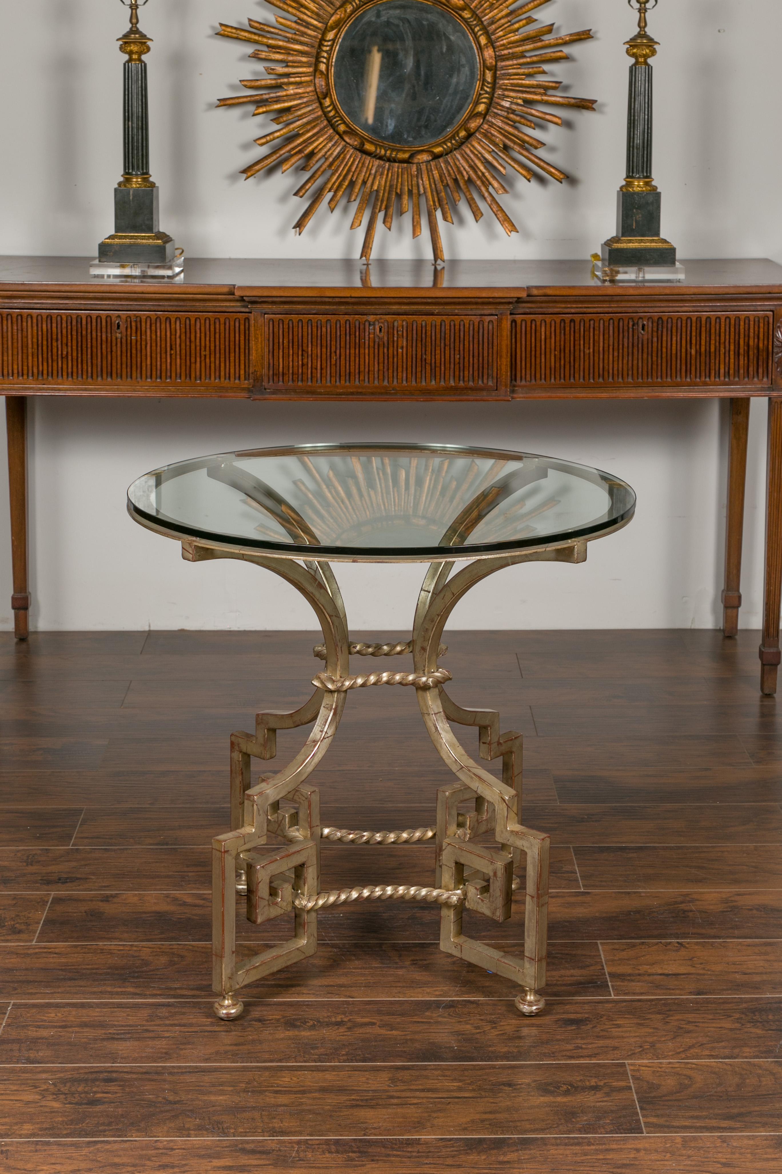A French vintage silver leaf iron center table from the mid-20th century, with circular glass top and Greek Key motifs. Created in France during the midcentury period, this table features a circular glass top resting above a silver leaf iron base