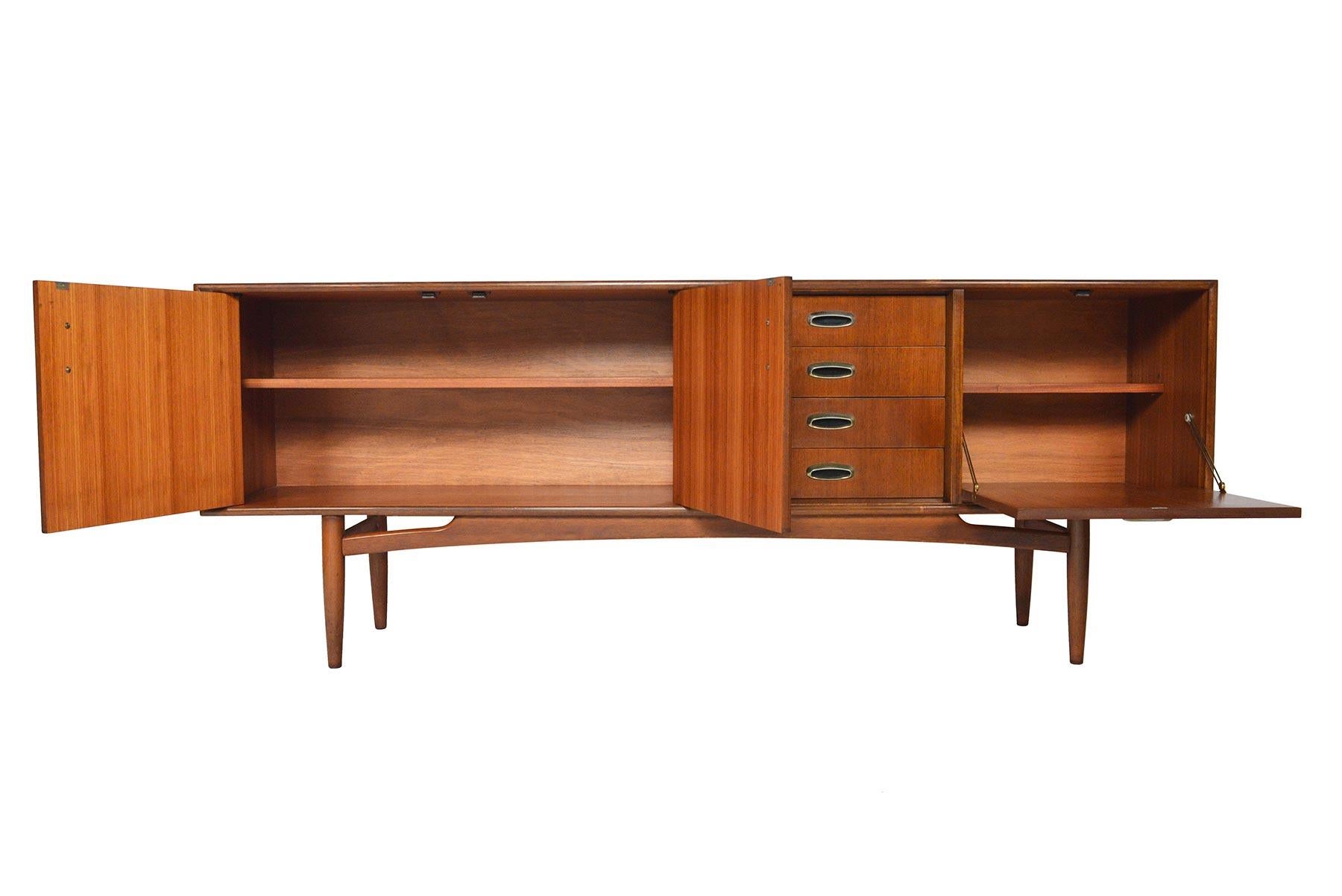 This English modern credenza is crafted in mahogany as part of G Plan’s Scandinavian range of the 1960s. A large left bay opens to reveal an adjustable shelf. A bank of four drawers sit to the left of drop down bar cabinet. Doors and drawers are