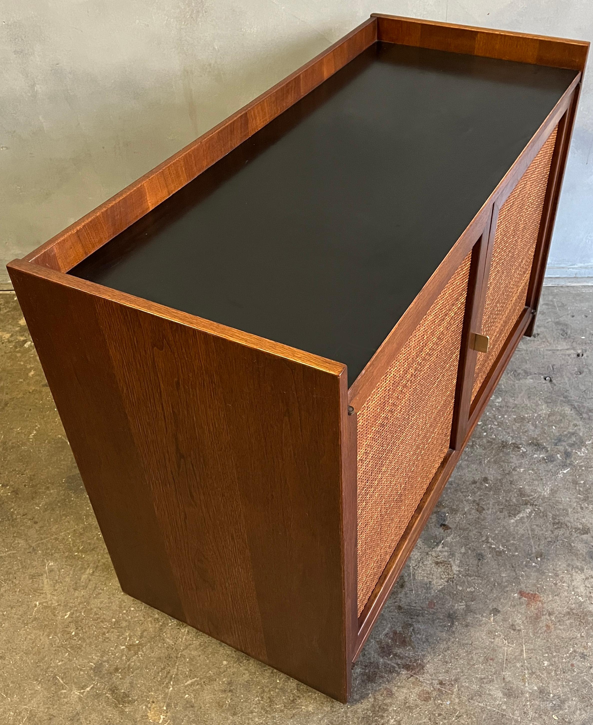 Midcentury double cane doors with gallery laminated top featuring two adjustable shelves and one pull our drawer. This item has a finished back and floating plinth base with hidden wheels on the underside. Superb quality walnut piece from the