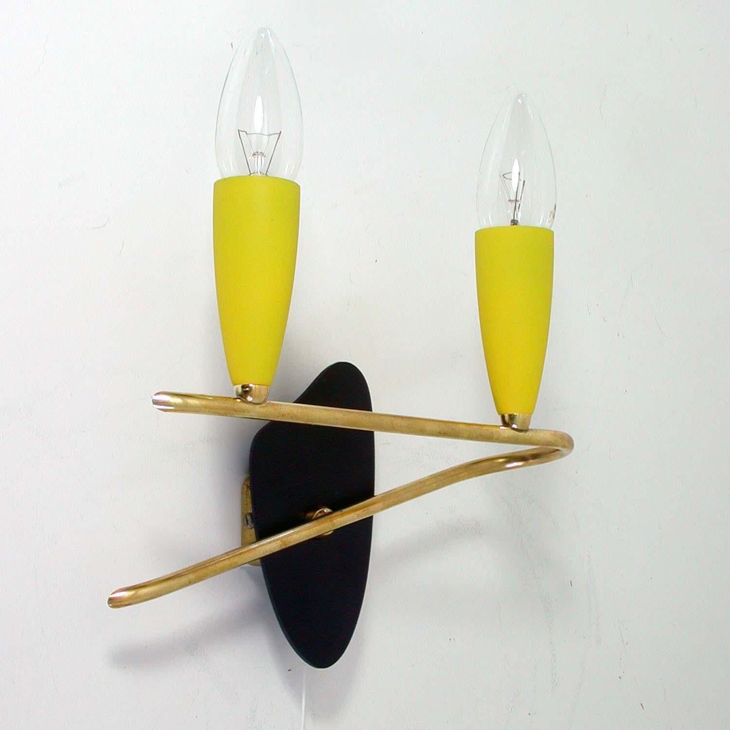 This cute little wall light was designed and manufactured in Germany in the 1950s. It is made of brass and has got 2 yellow lacquered E14 bulb holders. The backplate is black lacquered plastic in the shape of a painter palette. The switch for the