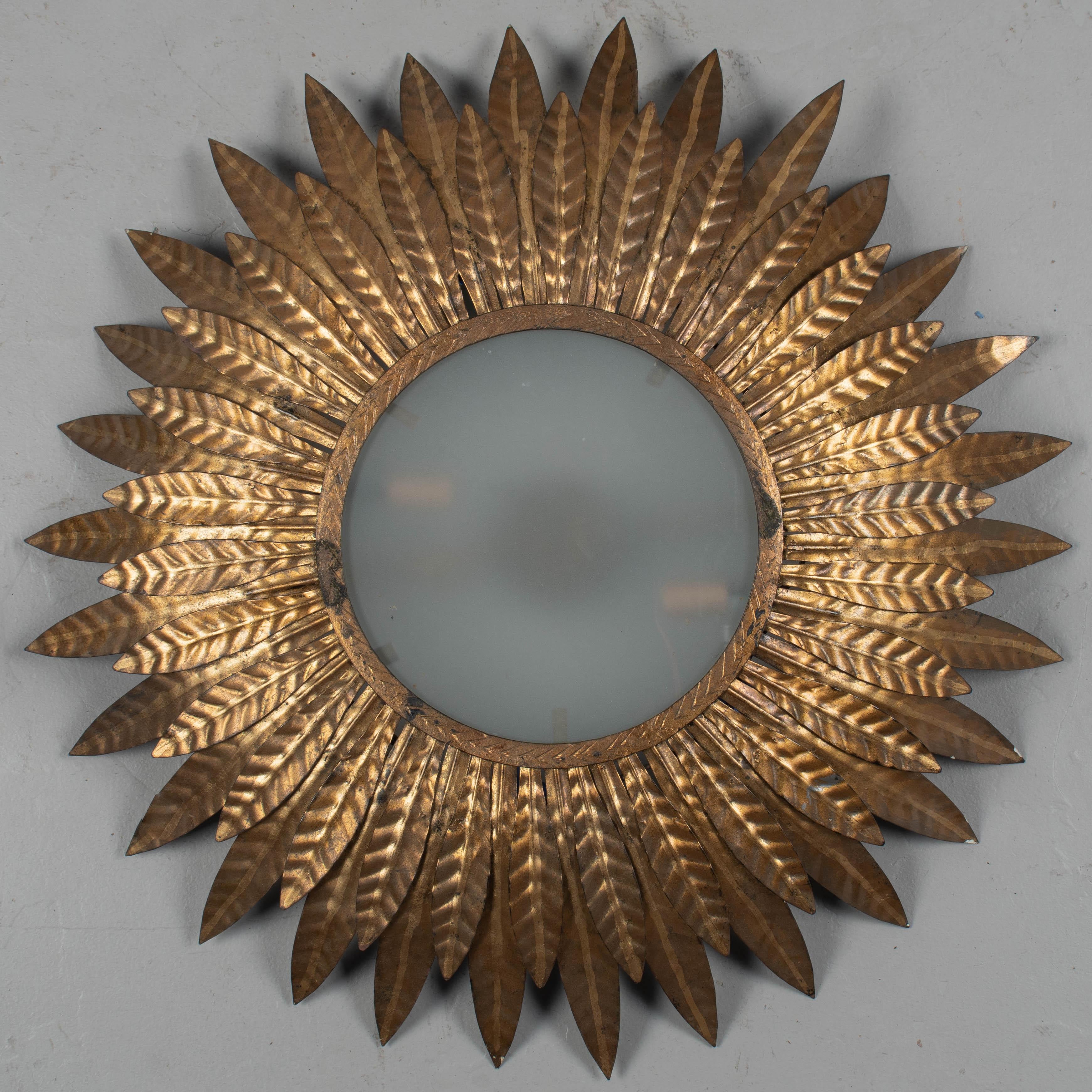 A Spanish Mid-Century Modern gilded metal sunburst flush mount ceiling fixture or wall light. Domed shape with two layers of wavy hammered tôle leaf form rays. Central frosted glass disc covers two light sockets. Beautiful effect when illuminated.