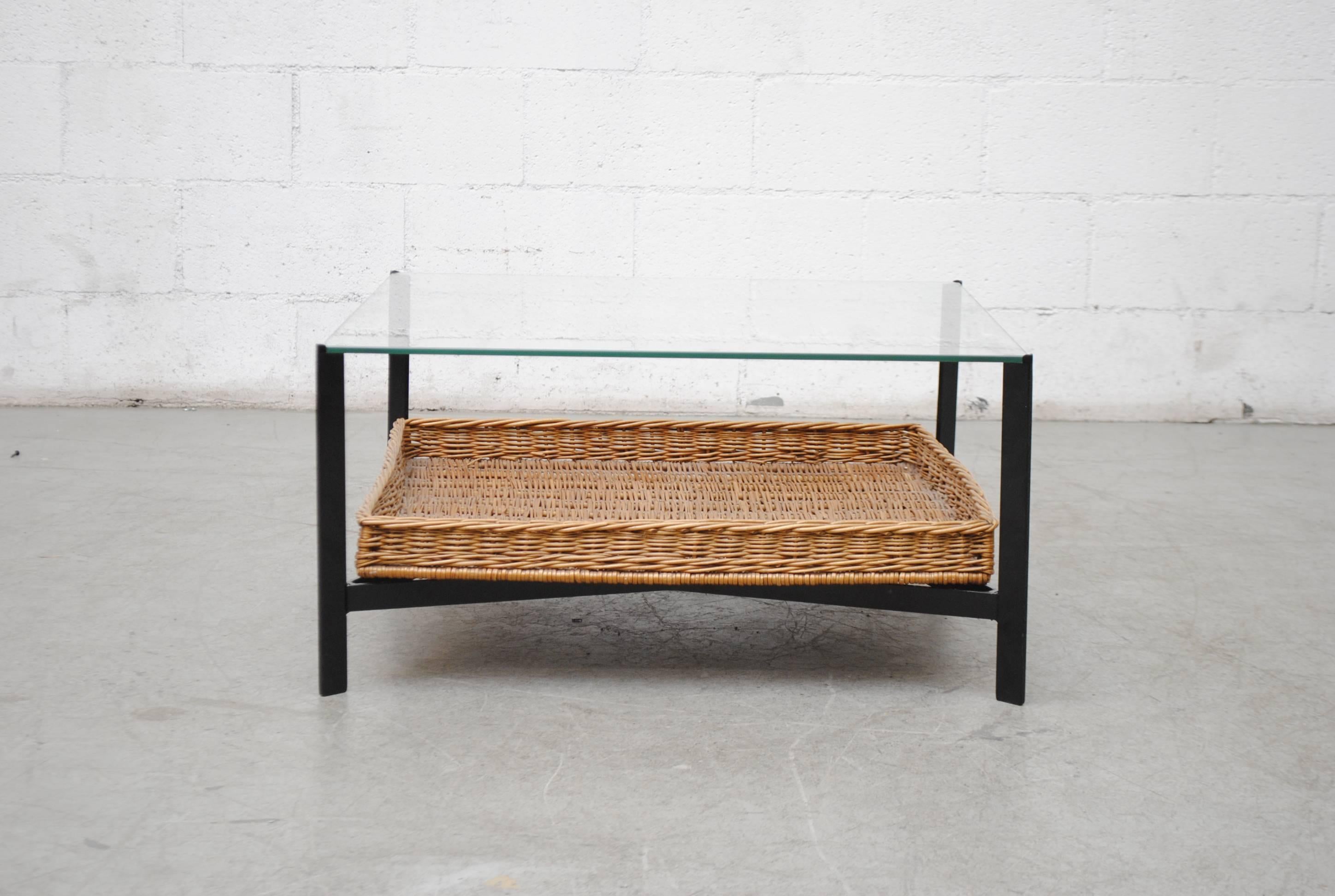 Square two-tiered glass-topped coffee table with woven rattan basket below. Black enameled metal frame with X-base. Good original condition. Visible wear consistent with its age and usage.