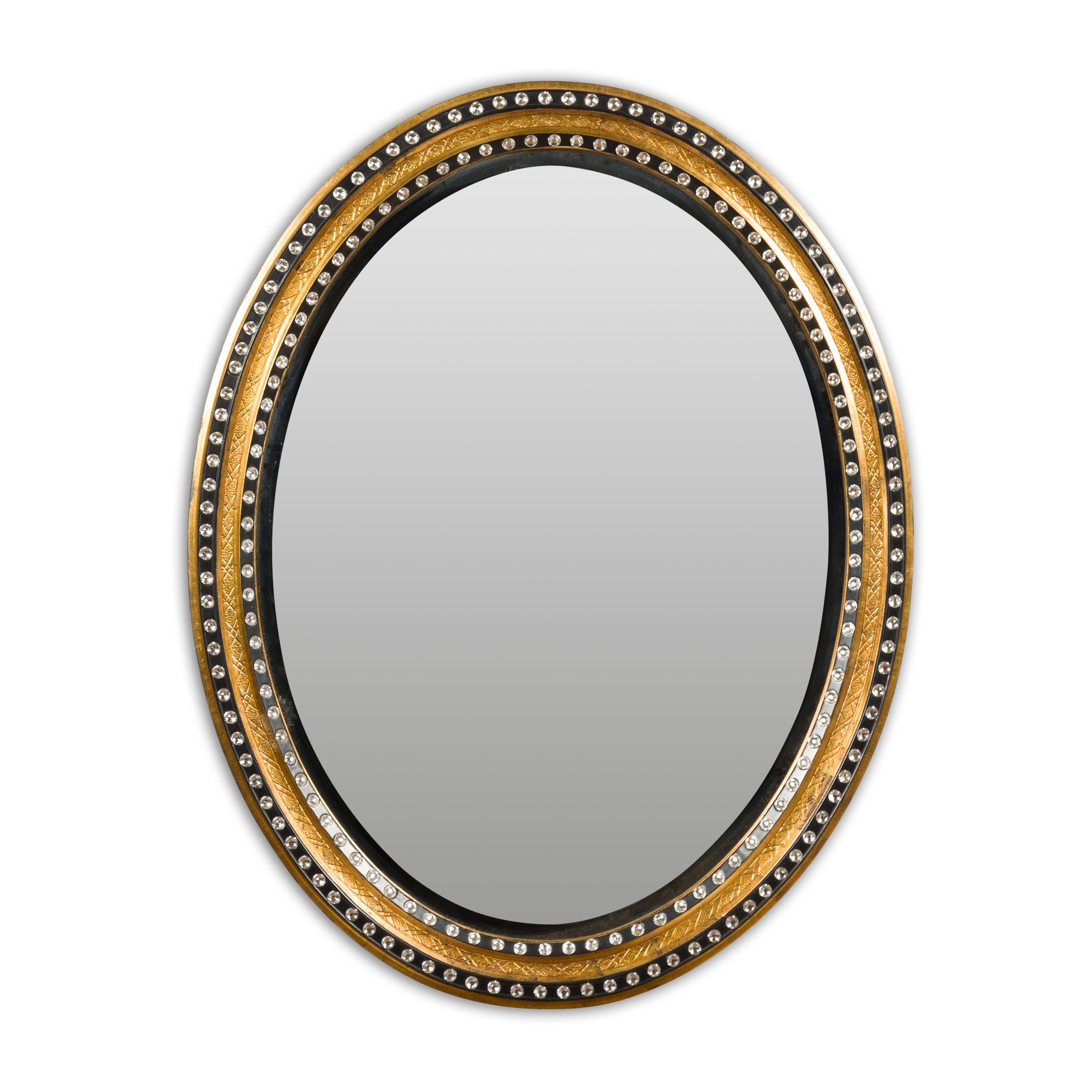An Irish Midcentury oval shaped mirror with gilded and ebonized wood, adorned with cut glass, diamonté décor. Embodying the eclectic charm of Midcentury design, this Irish oval-shaped mirror is an artful blend of elegance and whimsy. Framed in