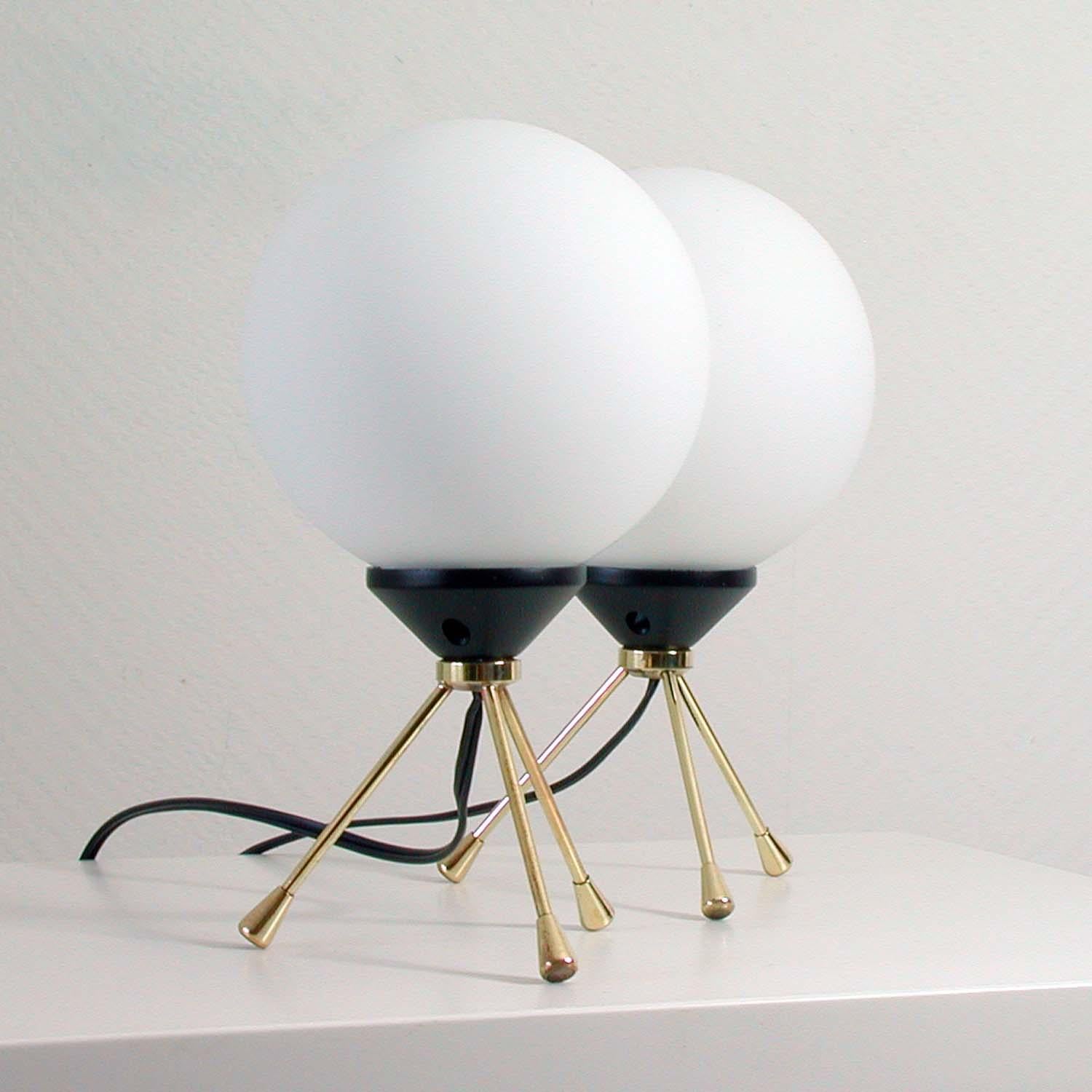 These 2 Sputnik style table lamps were manufactured in Italy in the 1950s. They are made of a brass tripod base with a black plastic bulb holder and white opal glass lampshades.