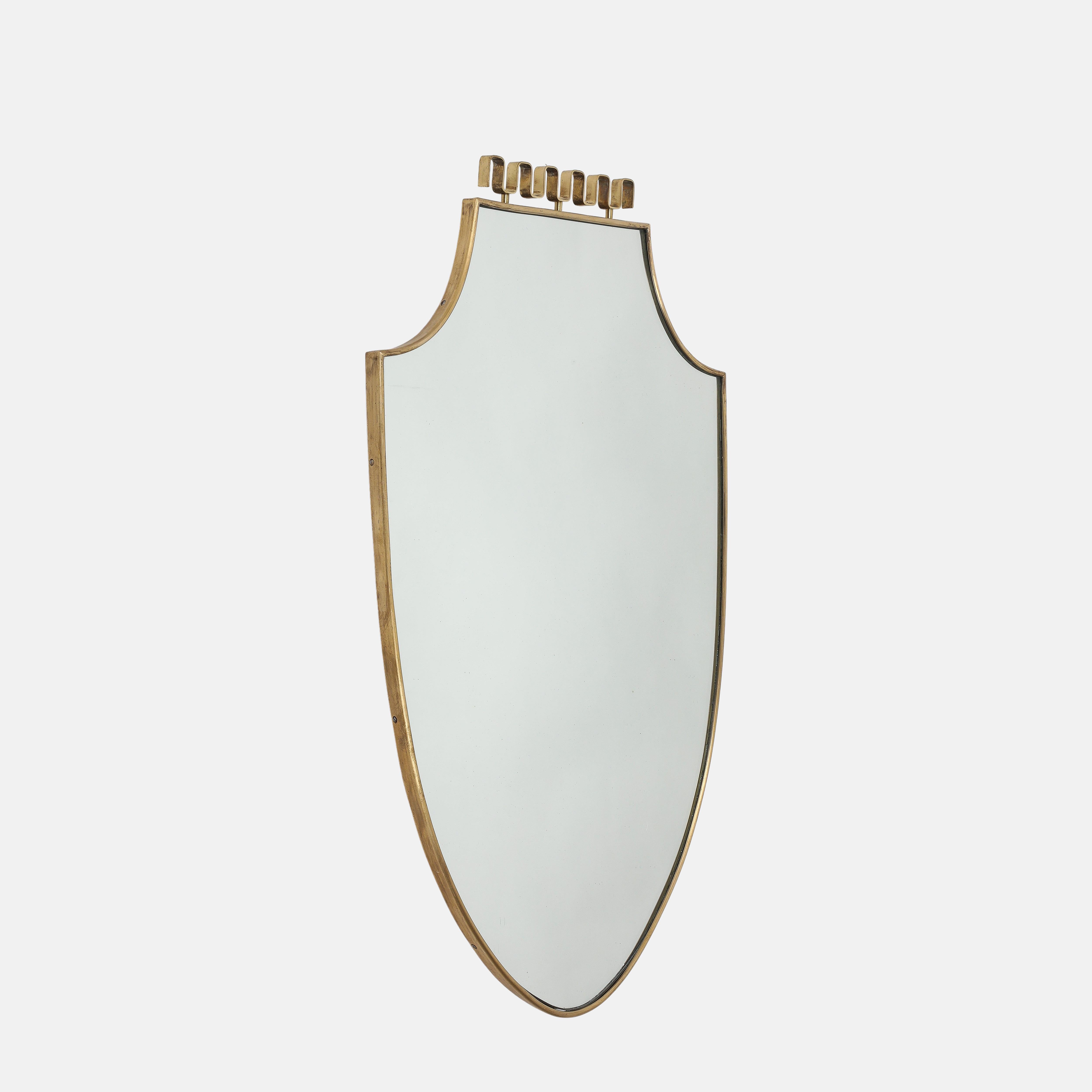 Espejo de pared italiano de latón en forma de escudo de mediados de siglo, compuesto por un marco de latón perfilado con la parte superior suavemente arqueada que se estrecha hacia la parte inferior, un precioso adorno central superior de latón en