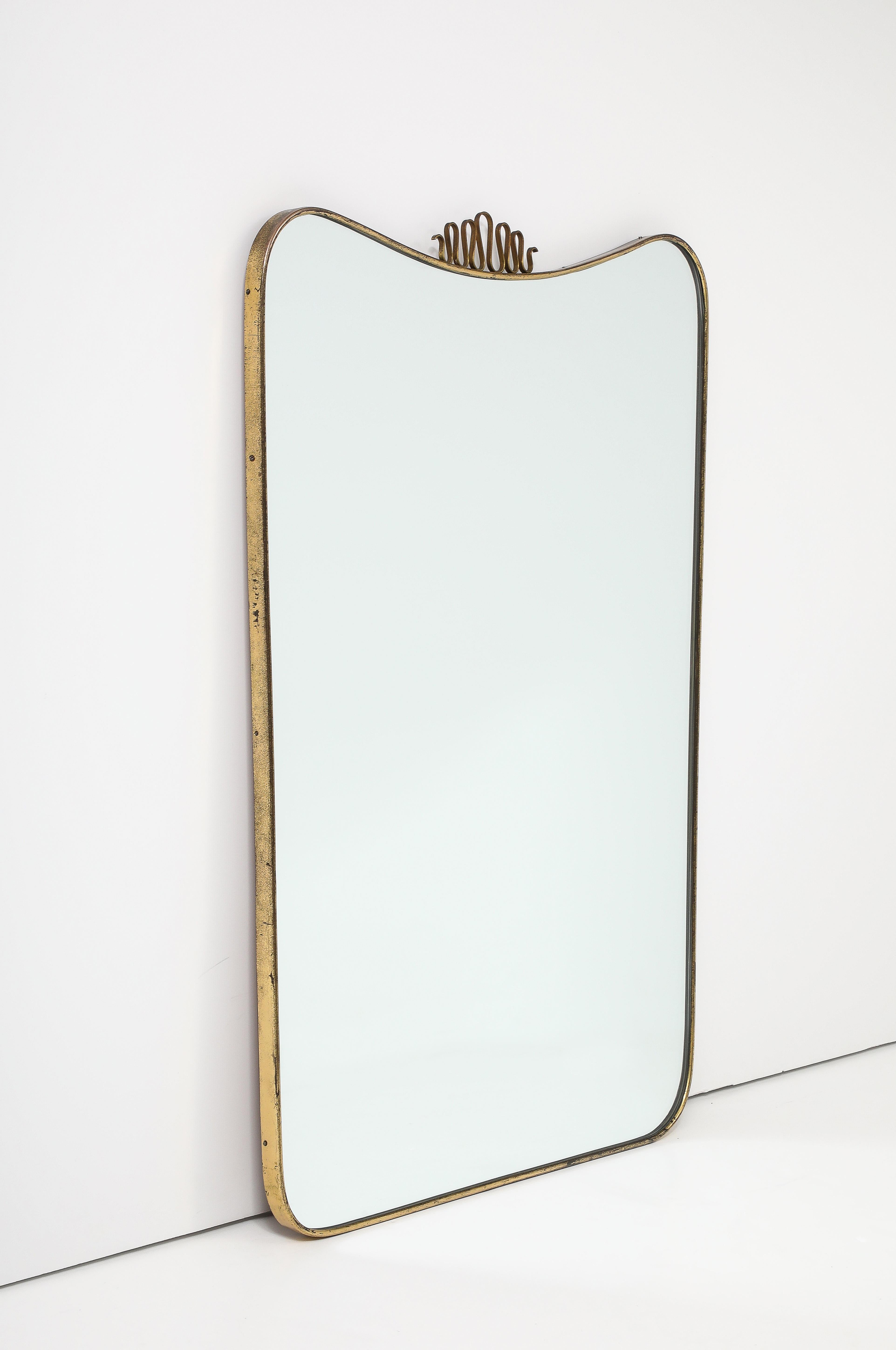 Midcentury Italian shaped brass mirror with central elegant decorative brass element on slightly arched and curved top slightly tapering down towards the bottom with rounded sides, in the style of Gio Ponti. This iconic and lyrical scroll top