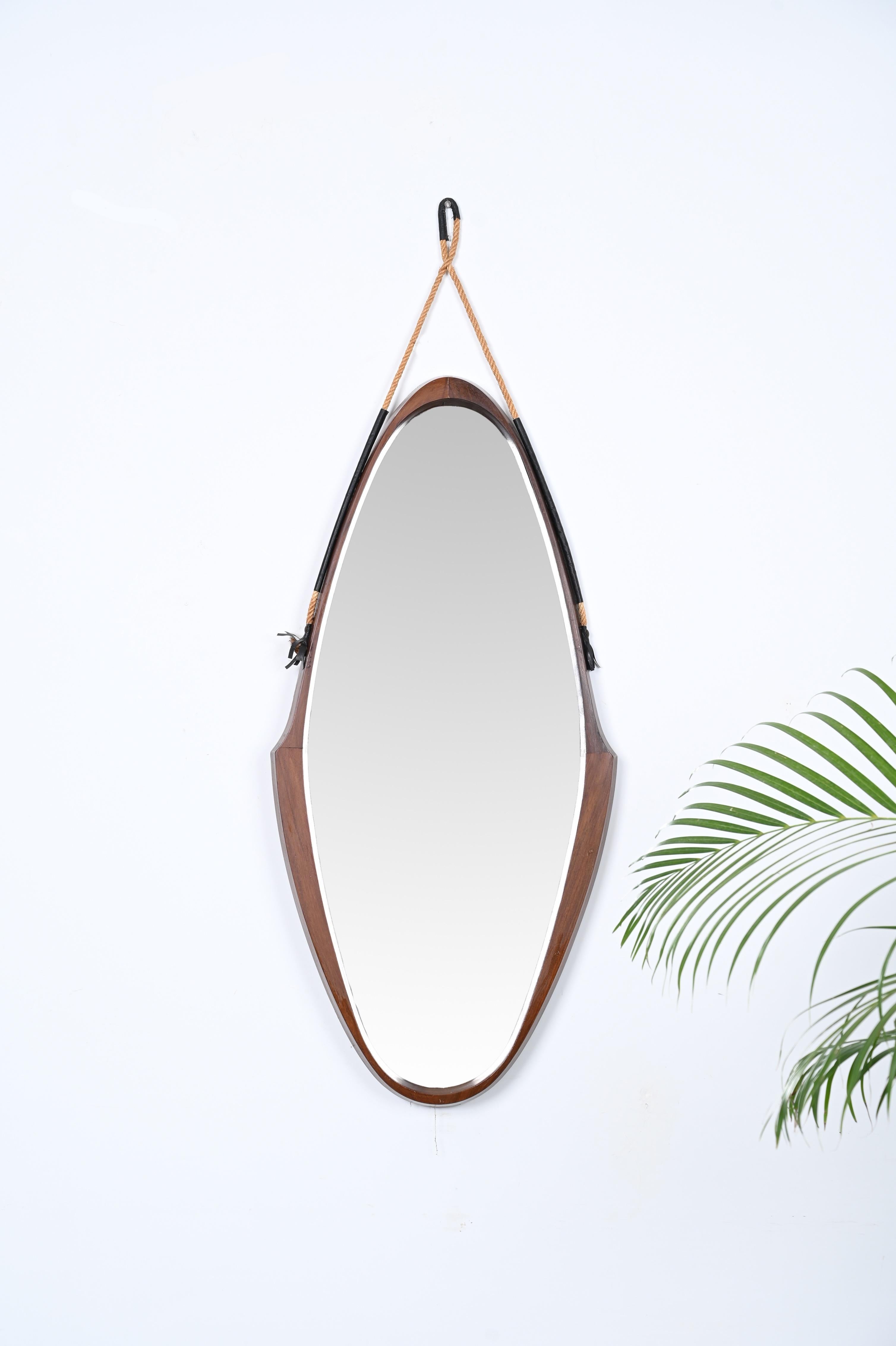 Stunning midcentury wall mirror designed in Italy during the 1960s. 

This elgant mirror features an astonishing oval shield-shaped frame made in a stunning curved teak wood, the structure seems to float thanks to the rope which is enriched at the