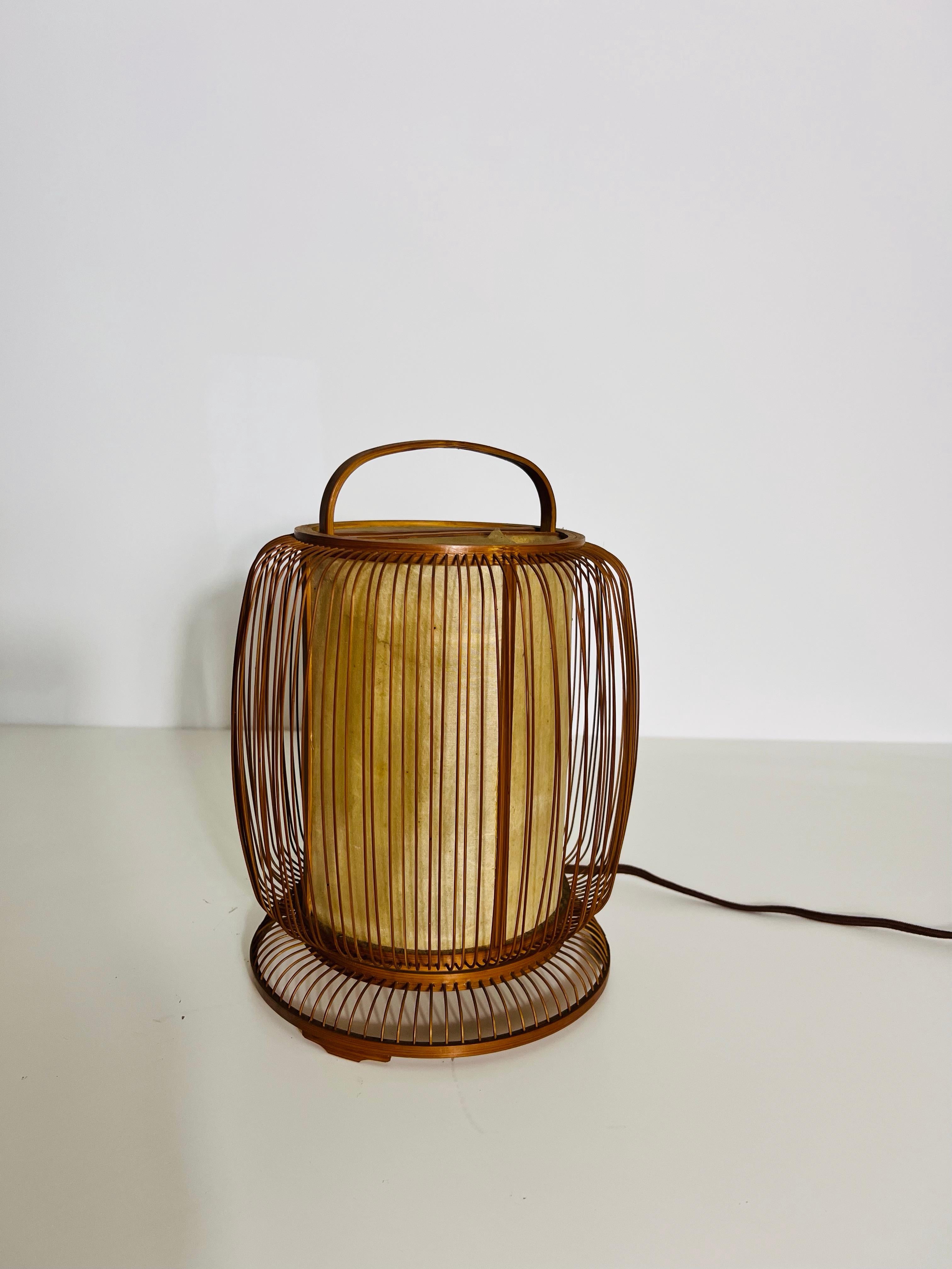 Cette délicate lampe de table du milieu du siècle capture l'essence du design japonais avec sa construction en bambou naturel et son élégance discrète. Fabriquée à partir de fines tiges verticales de bambou entourant un abat-jour intérieur, cette