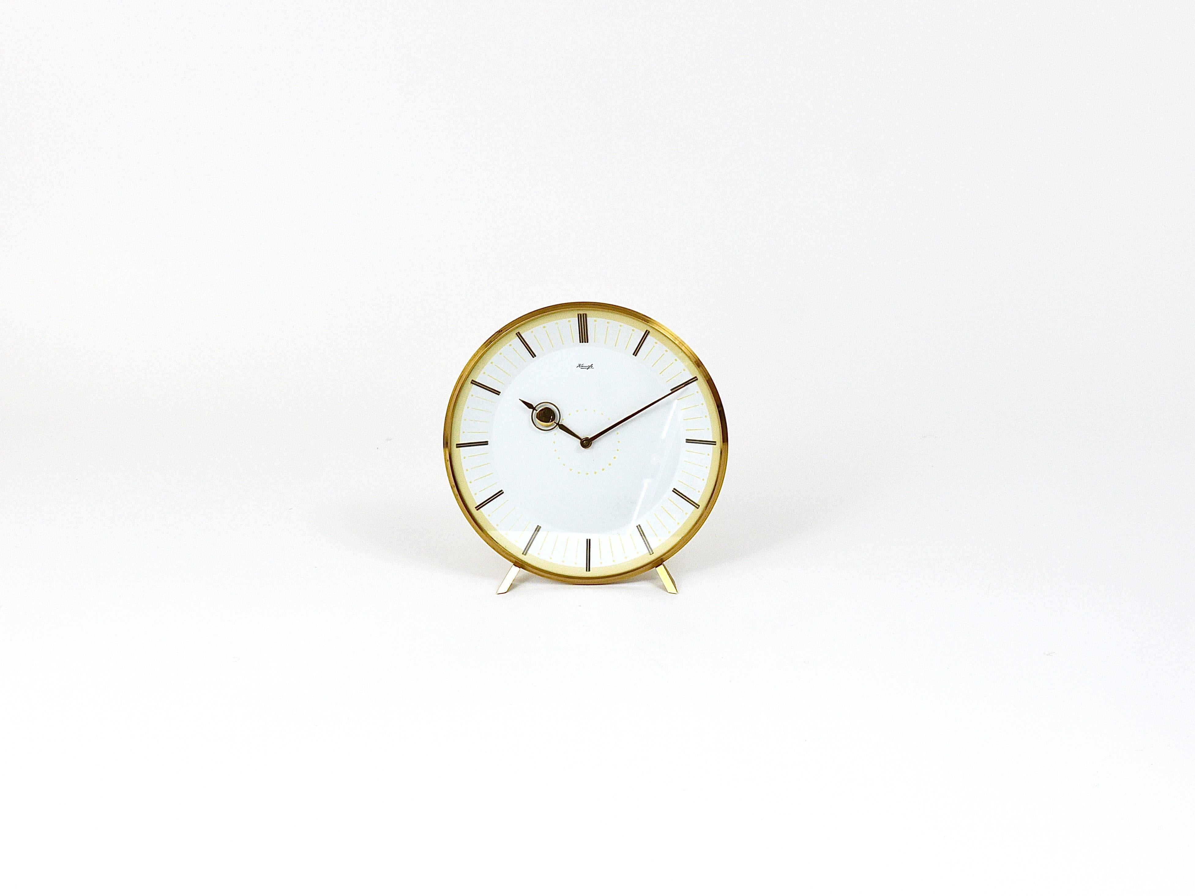 An elegant Mid-Century Modern desk or table clock with a mechanical 8-days movement from the 1950s. Executed by Kienzle Germany. This charming clock has a partly polished brass housing, a lovely clocks face with pastel yellow and golden indices and