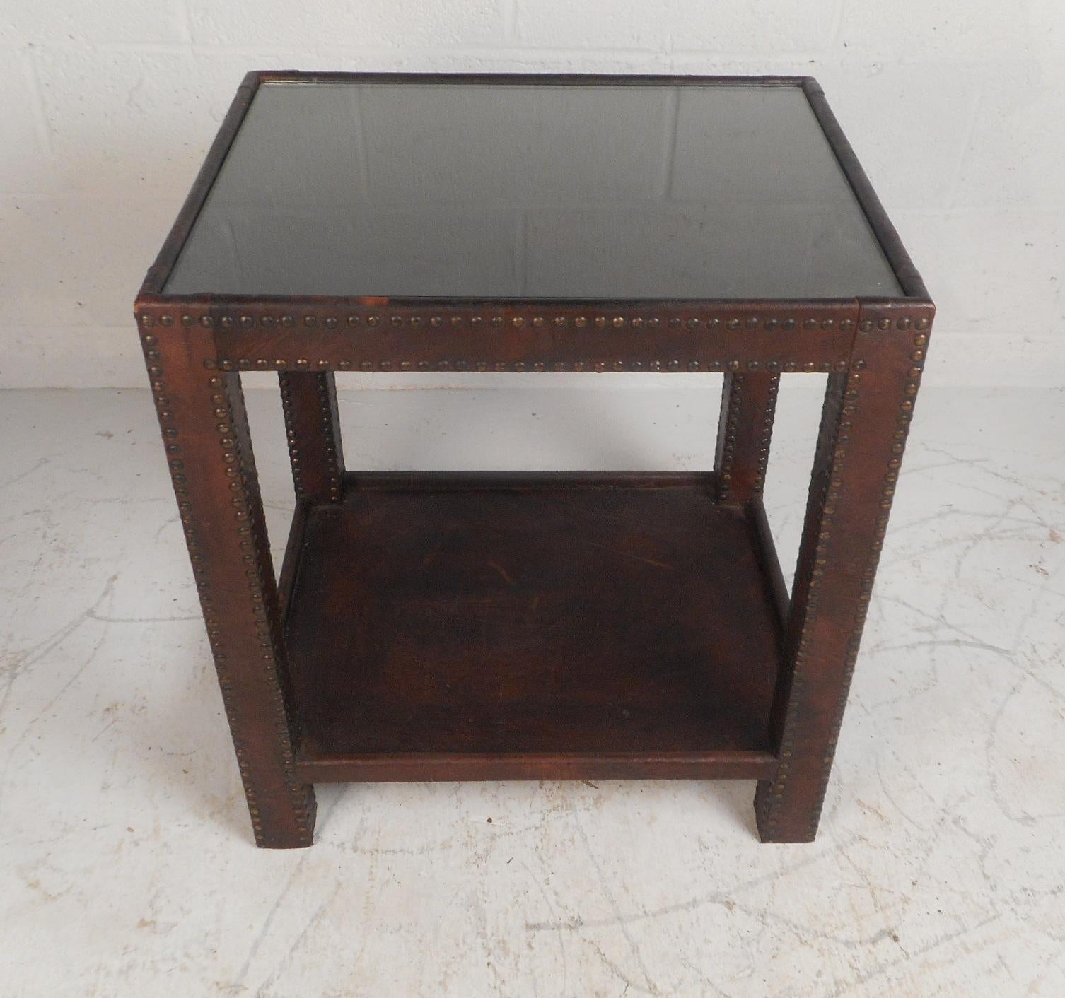 This beautiful vintage modern two-tier side table is covered in brown leather with studs running along the legs and front. A one of a kind piece with a glass top and a lower shelf for additional storage. This lovely midcentury pedestal table makes