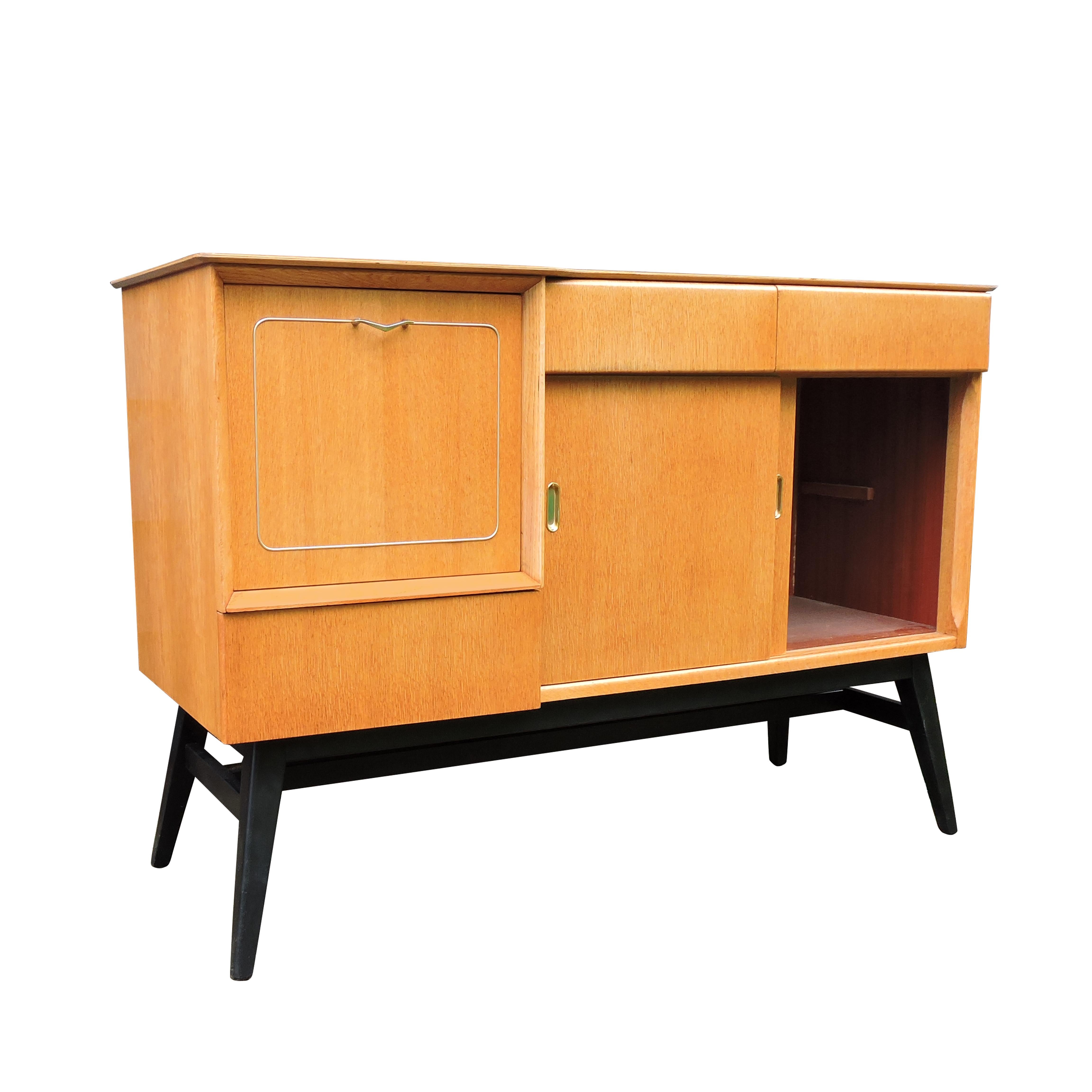 A midcentury light oak sideboard with upper drawers, a flip down cabinet, and sliding doors. It features fine brass detailing and tapered black legs.