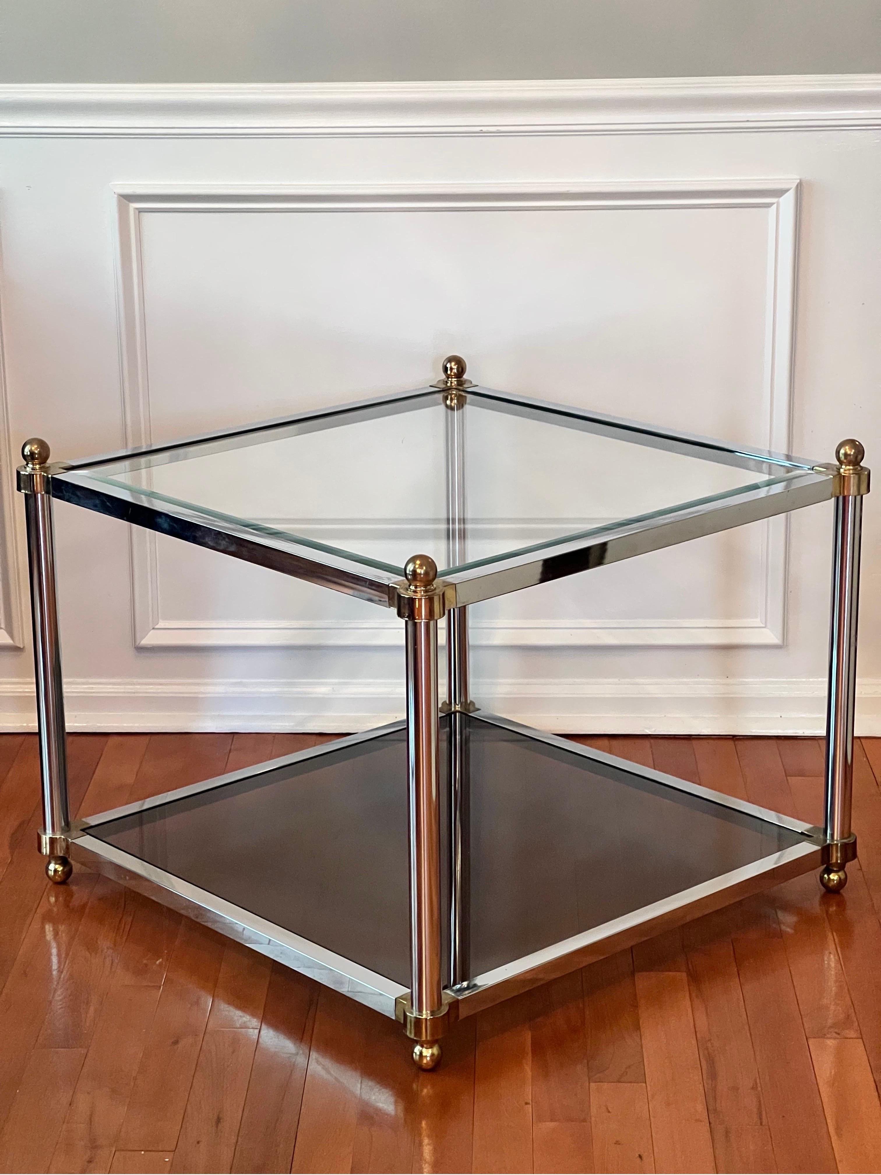 Elegant 1970's two tier brass and polished steel square cocktail/coffee table in the manner of Maison Jansen. 

The lower tier features smoked glass while the upper tier is beveled, transparent glass. Brass corners and ball finials adorn the table