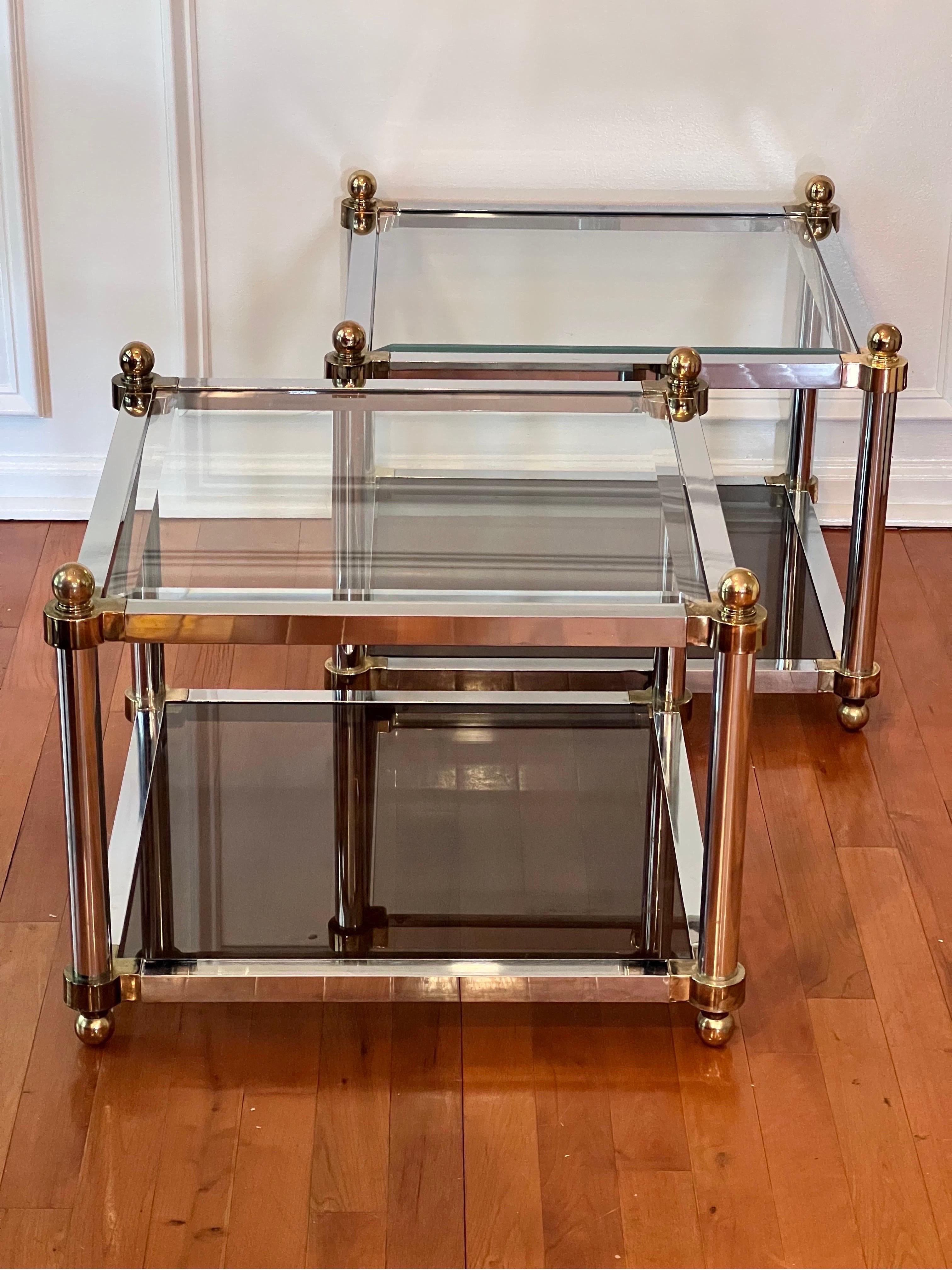 Handsome pair of 1970's two tier brass and chromed polished steel square tables in the manner of Maison Jansen. 

The lower tier features smoked glass and the upper tier is transparent, beveled glass. Brass corners and ball finials beautifully adorn