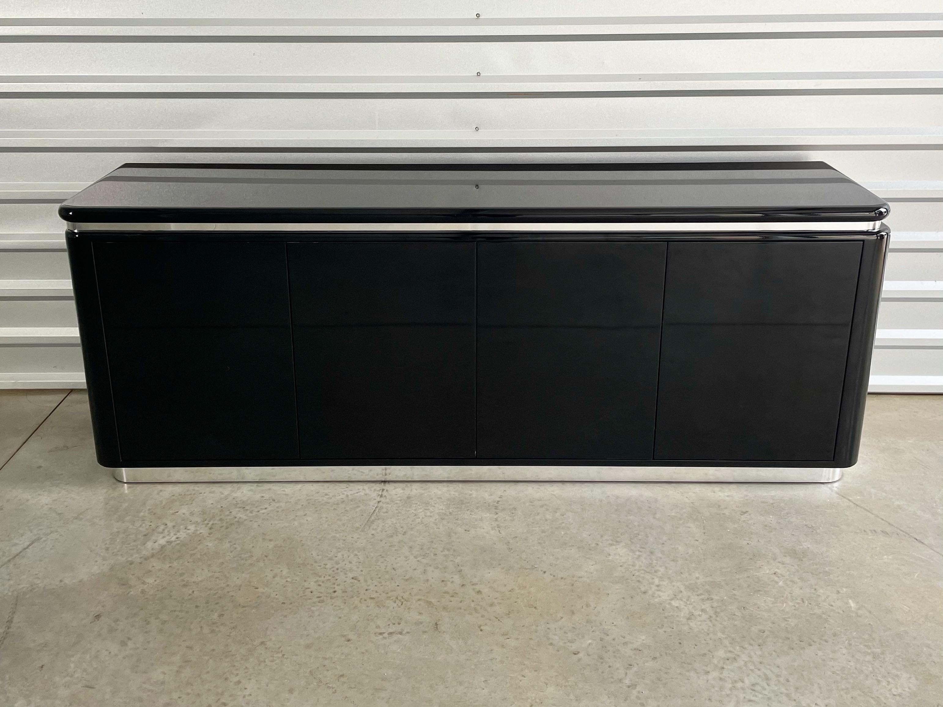 Black lacquer and chrome modernist credenza in the manner of Milo Baughman, circa 1970s. Exquisite design and craftsmanship. Two sets of double cabinet doors open to reveal drawers and shelved storage. Black and white interior. Chrome banding detail