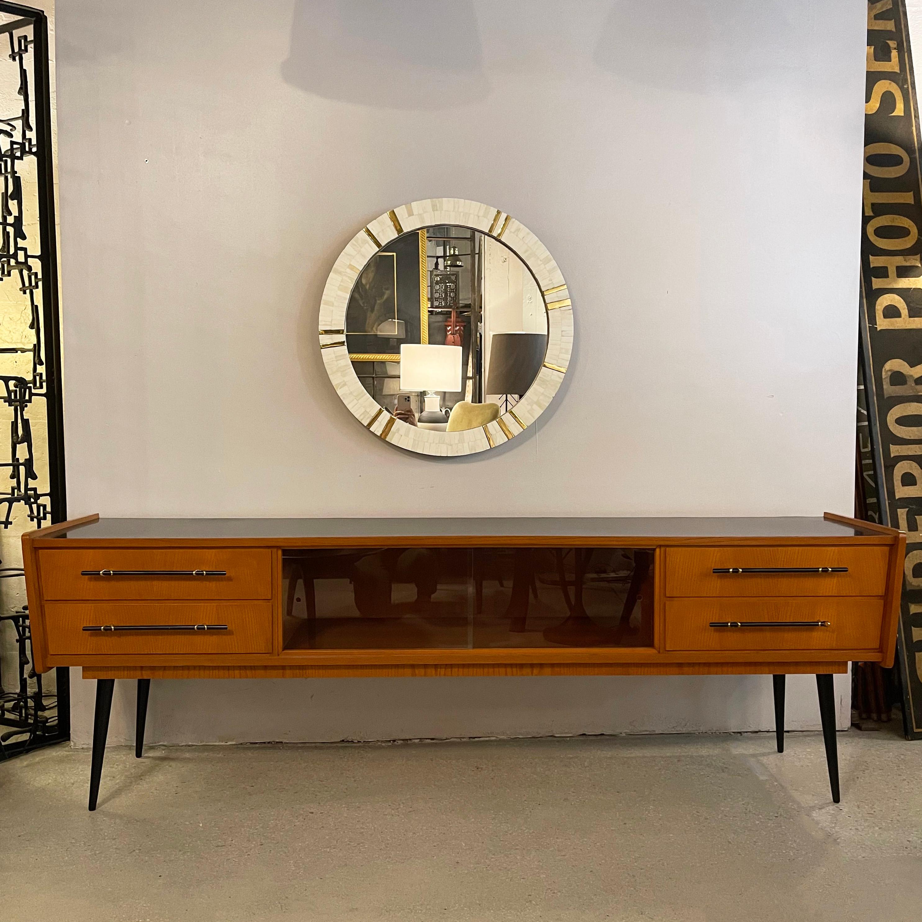 Mid-century modern, long and low, ash wood, console credenza features contrasting, inset black laminate top, black tapered legs and black bar handles with wrapped brass hardware on it's four slim drawers. The center has sliding glass doors for