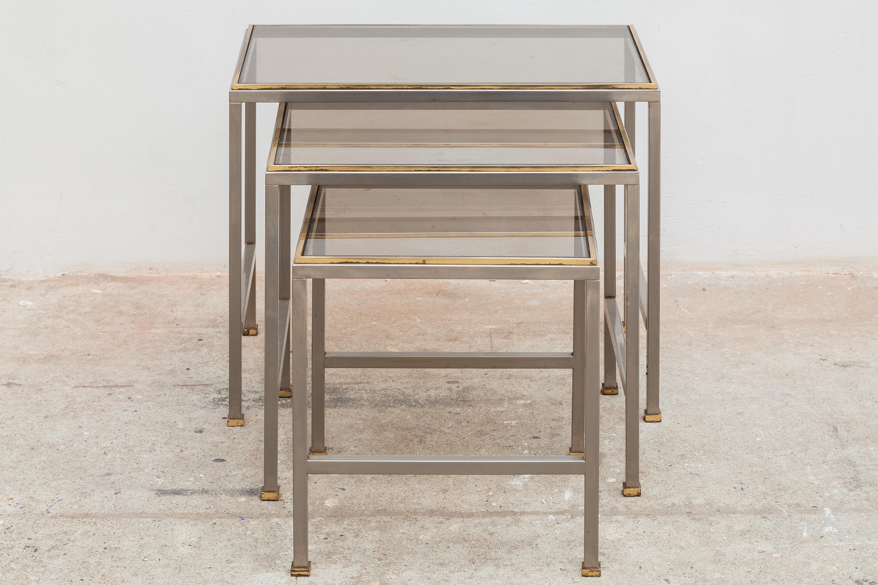 Vintage mid-century nesting tables. Modernist silver and brass tone frames with smoked glass tops.Dimensions:Small:32W x 34H x 32D cm Med: 42W x 38H x 32D cm Large: 52W x 42H x 32D cm.