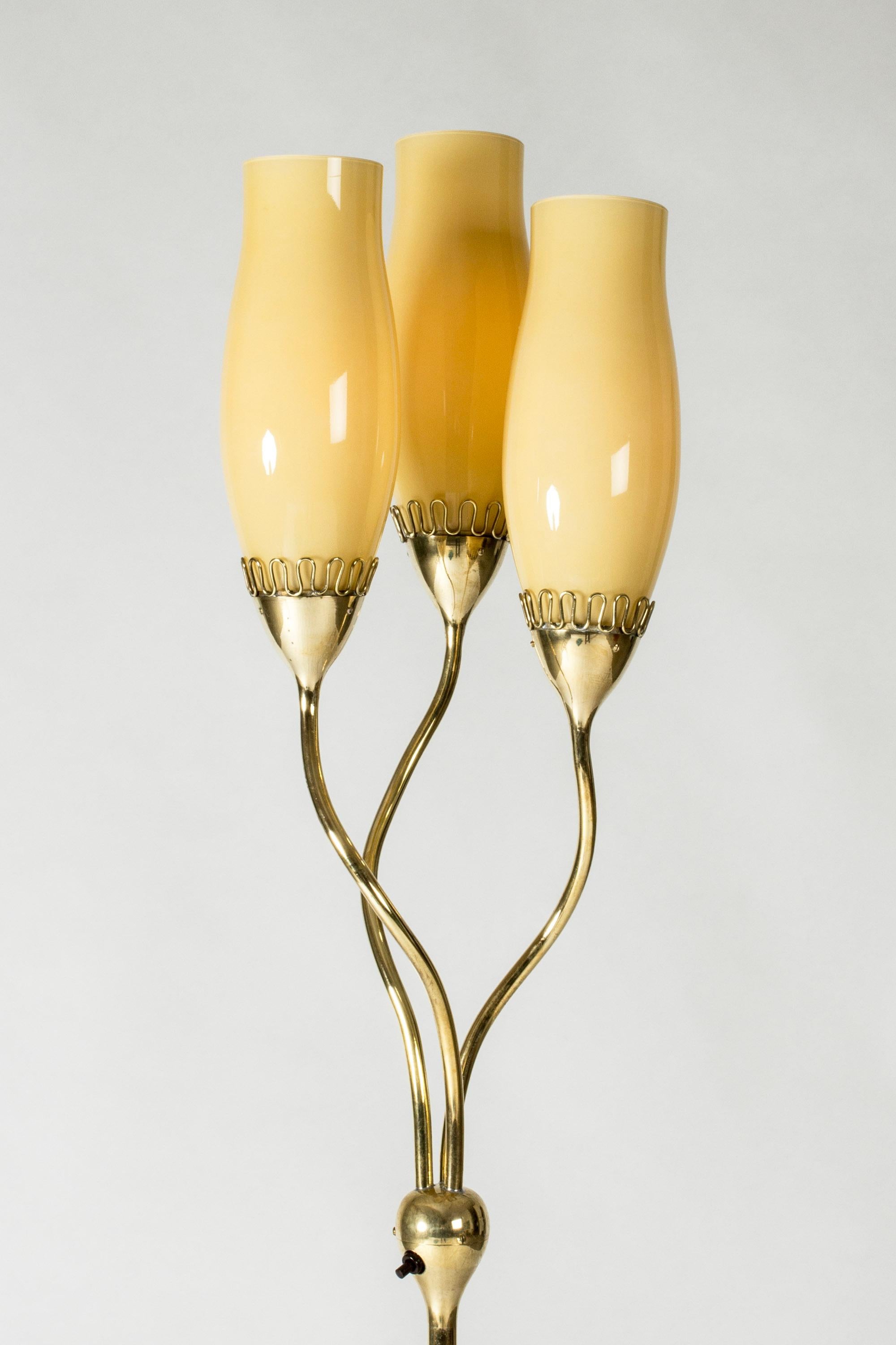 Preciosa lámpara de pie de Mauri Almari, de elegante diseño, con tija de latón rematada en tres brazos ondulantes que sostienen pantallas de cristal amarillo. Expresión orgánica, las sombras proyectan un brillo cálido.
