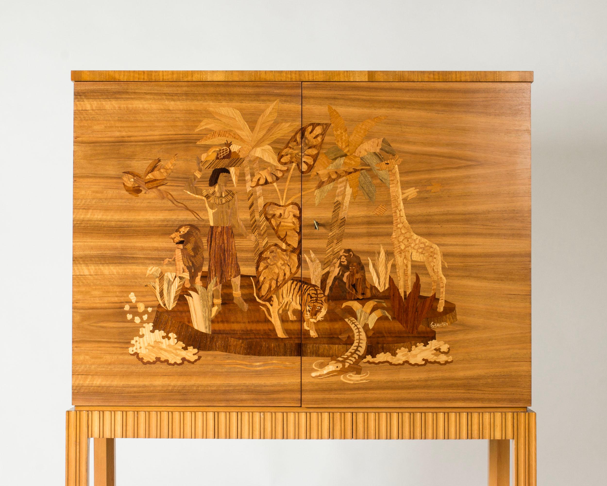 Magnifique meuble de Mjölby Intarsia, fabriqué en acajou avec des portes incrustées d'un motif paradisiaque par Erik Matsson. Elle représente un homme et des animaux sur une petite île, soigneusement rendus avec du bois dans différentes