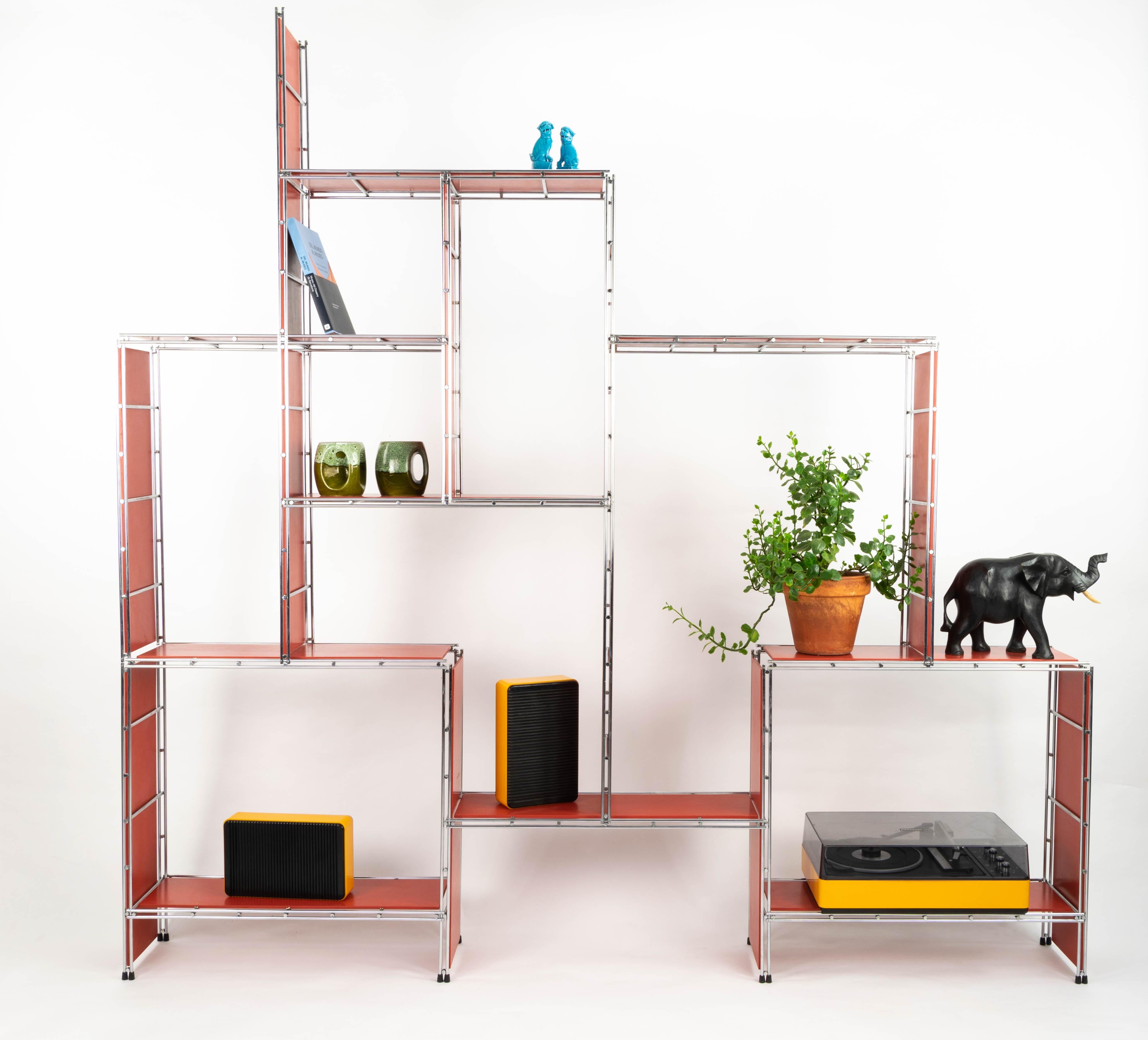 Etagère composée de 20 pièces de structure en métal chromé et bois laqué rouge qui permet de multiples montages grâce à un système sans vis breveté par la marque. Possibilité de montage comme étagère au sol ou au mur. Il est conçu de manière