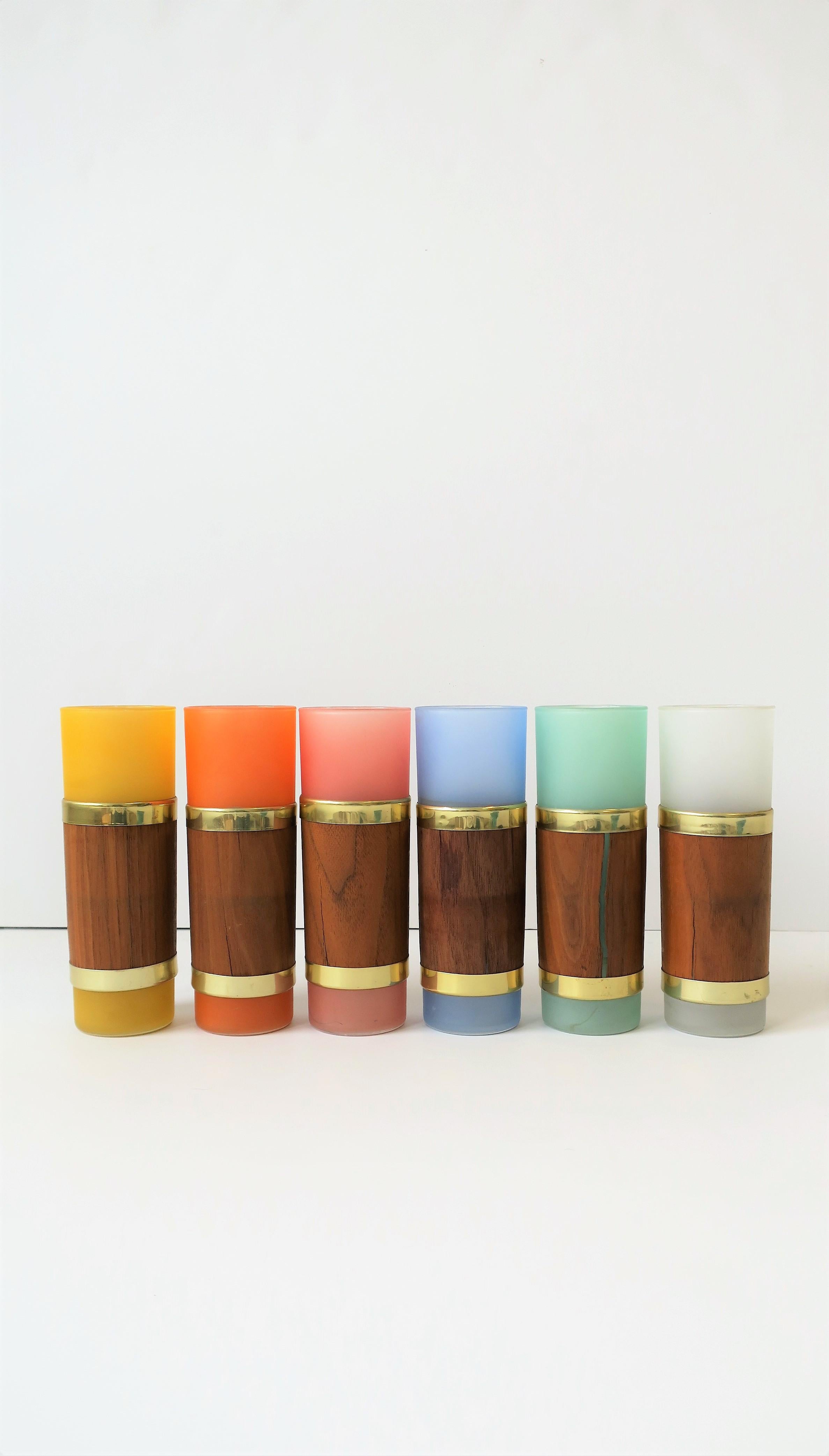 A chic and rare set of six (6) Mid-Century Modern colorful highball cocktail or beverage glasses with wood and brass detail, circa 1960s. A rainbow of colorful 'frosted' glassware wrapped in wood and gold-tone metal, from left to right: Yellow, red,