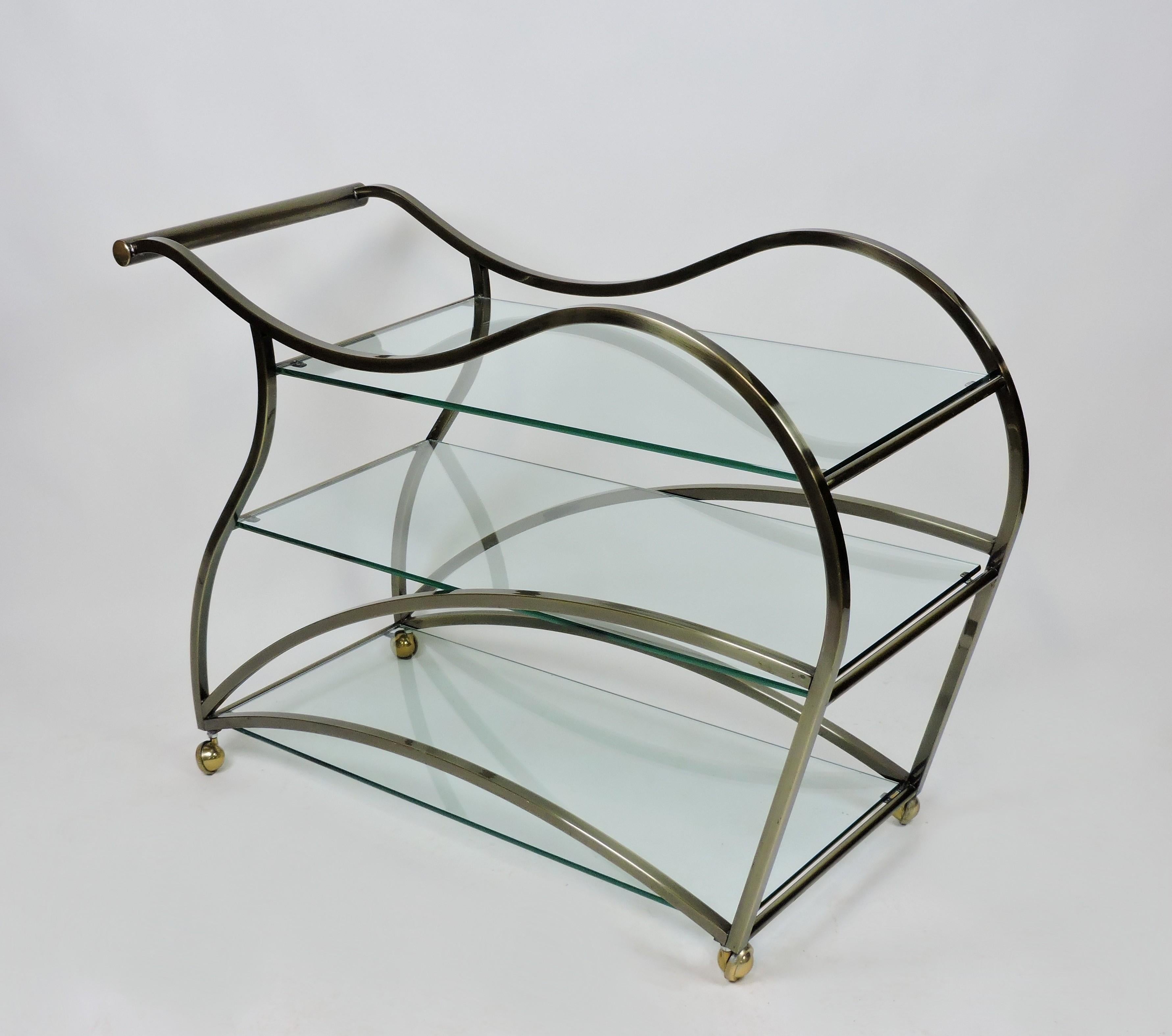 Sexy and curvaceous sculptural bar or tea cart manufactured by high quality furniture maker, Design Institute of America. This rolling cart has a metal frame with an antiqued brass finish and three, 3/8 inch thick, clear glass shelves. Its large