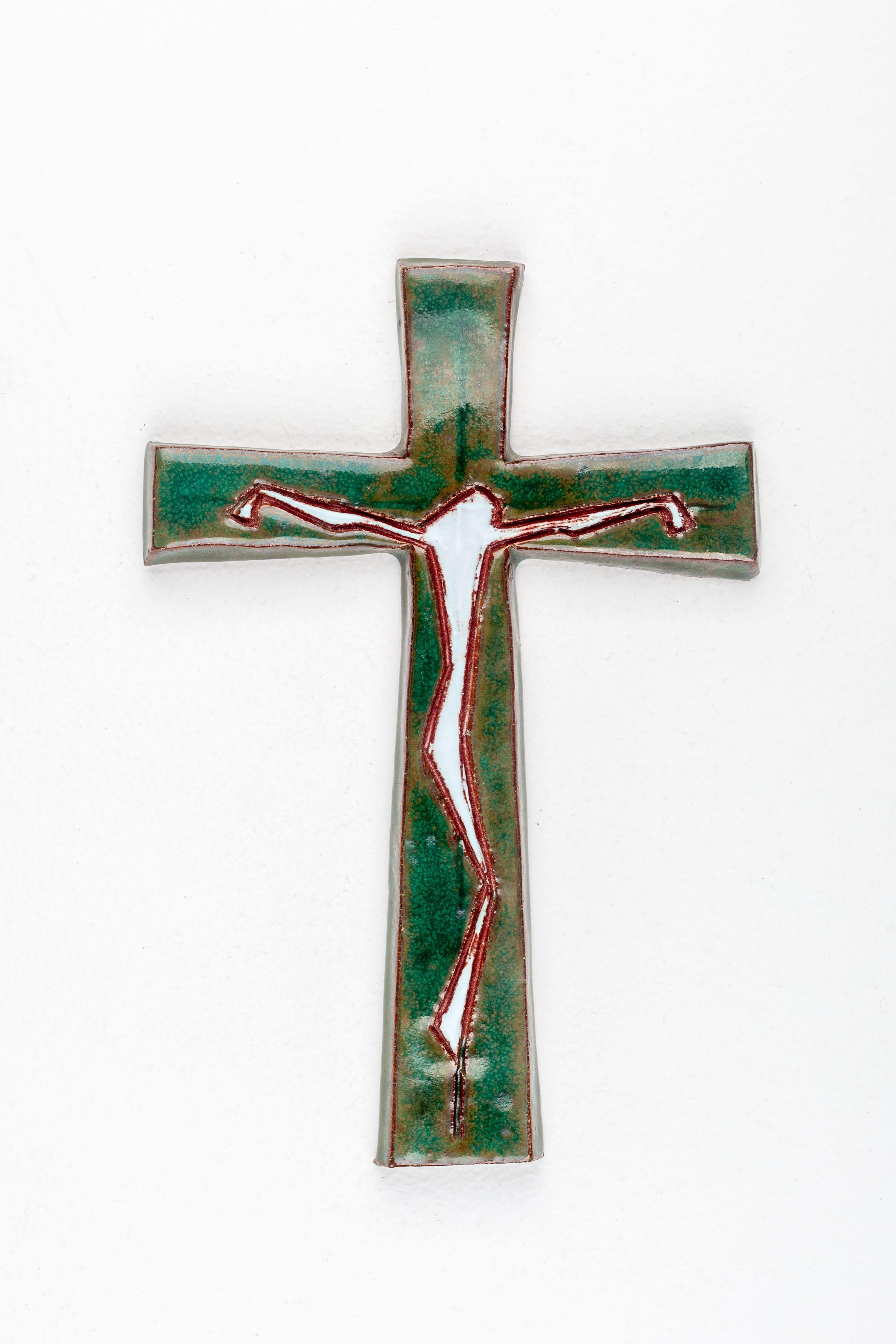 This ceramic wall cross, hand-painted in textured greens and ocher beige, features a depiction of Christ in painted white. The angular portrayal of Christ, rendered in straight terracotta lines, stands at the center of a cross with larger ends. This