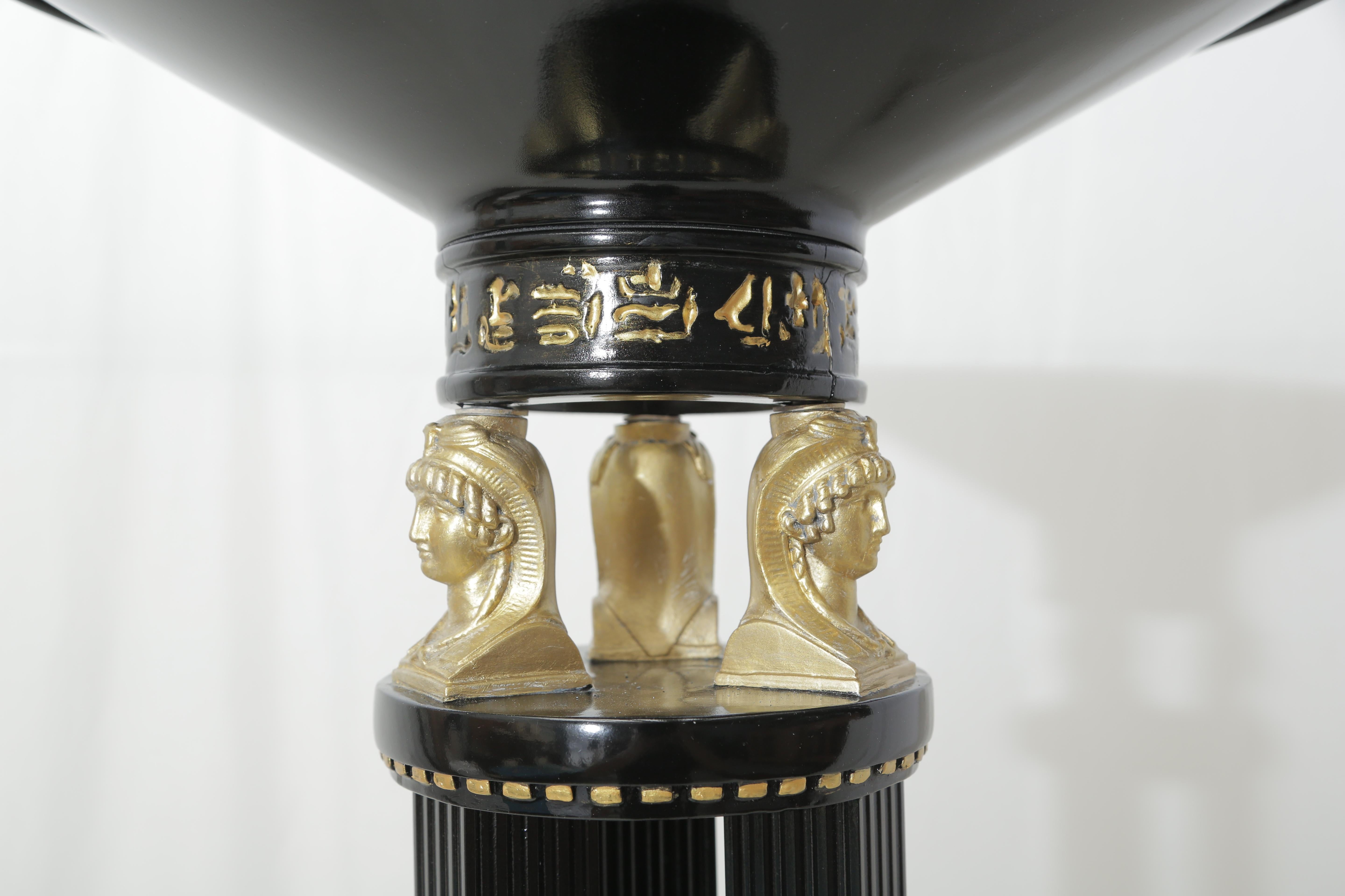 Black lacquered metal floor lamp with gold leaves Egyptian figures supporting the top.
Egyptian signs around the base of the top.
 