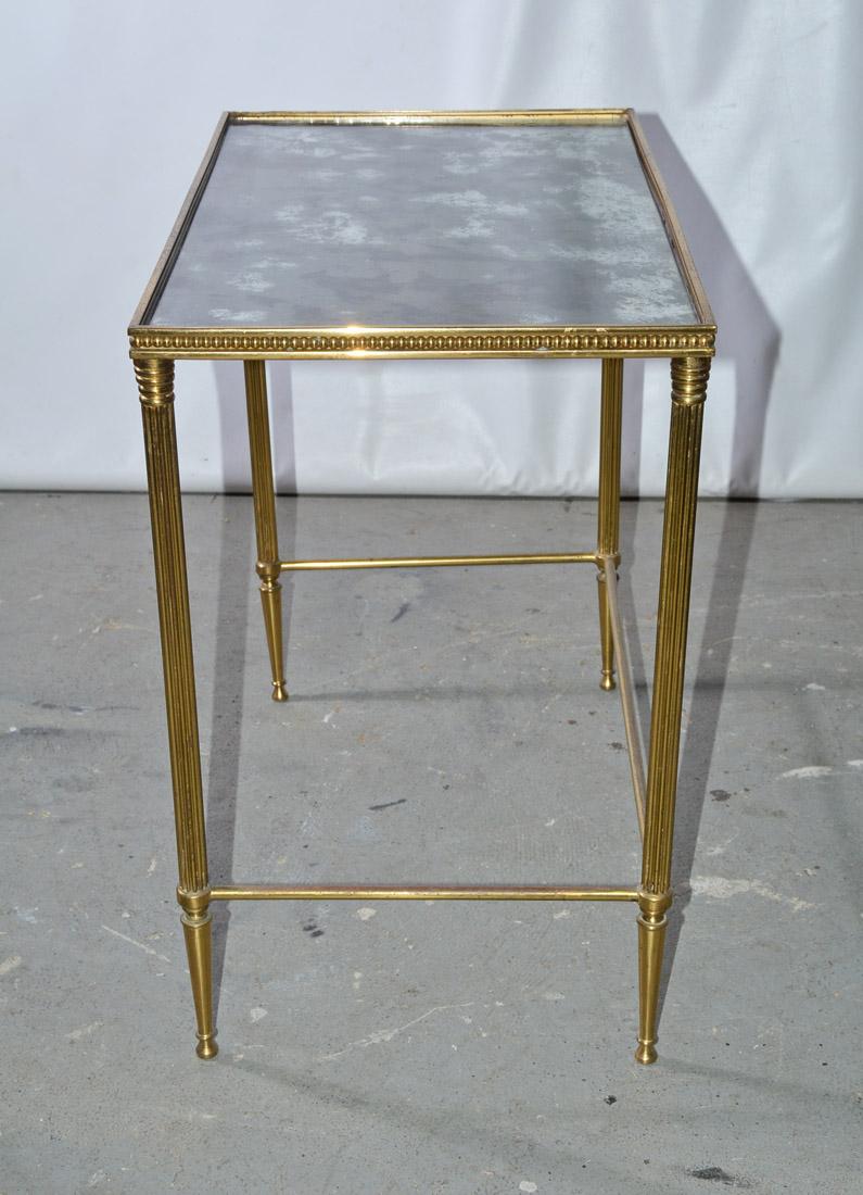 The midcentury neoclassical Maison Jansen style brass side or end table has a smoky mirrored top framed with a beaded edging, fluted legs and tapered feet. There are brass stretchers on three sides so that the table can be pulled up to a seated