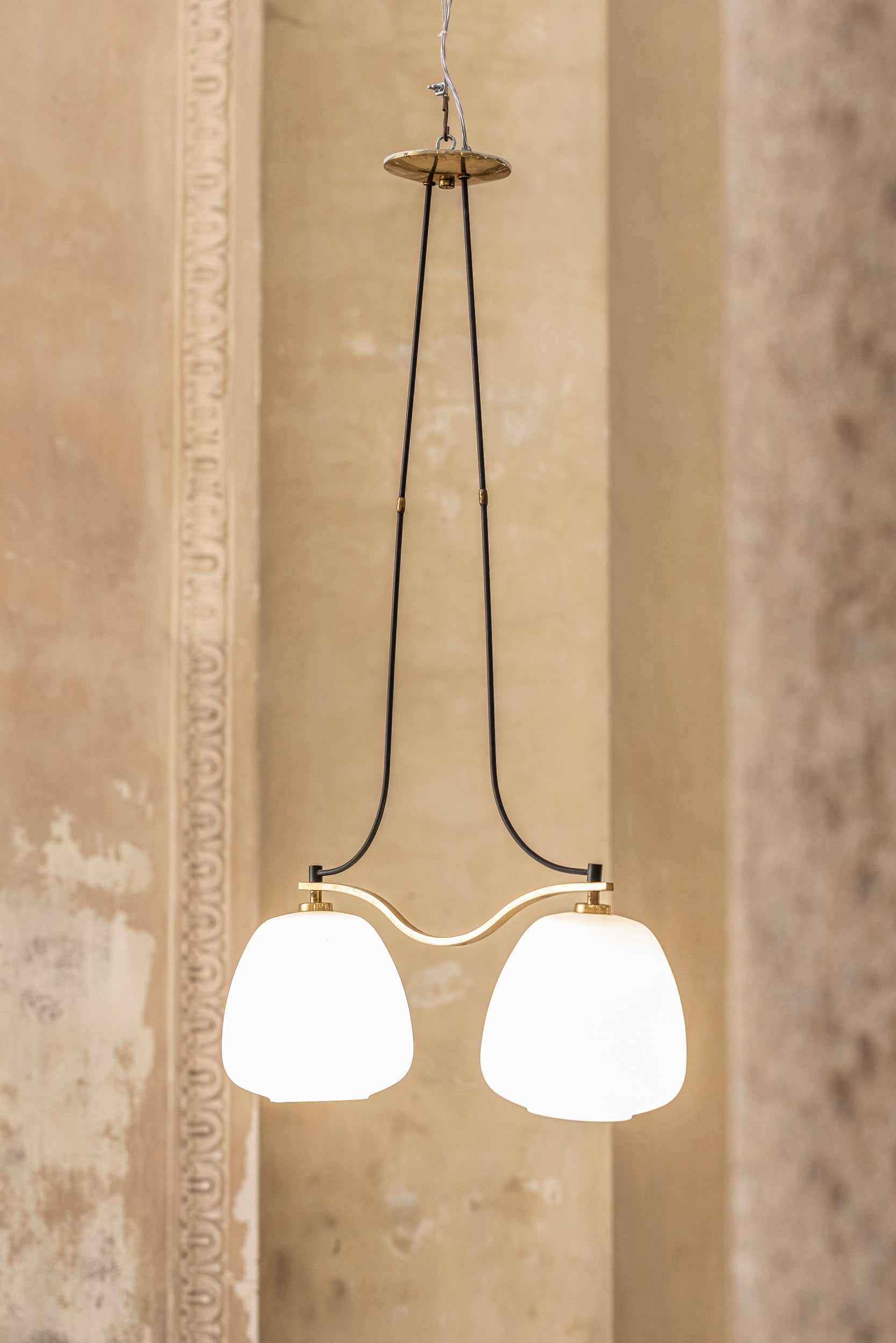 Elegante lampadario di metà secolo con una delicata struttura in ottone dorato e due paralumi in vetro opalino, finemente lavorati e decorati. 
Questo ciondolo di manifattura italiana degli anni '50 combina un design raffinato con una sobria