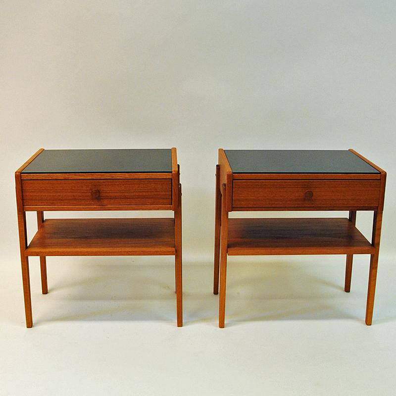 Lovely and Classic pair of vintage teak and glass top bedside tables with drawers and straight legs. These midcentury night tables has a black painted shiny glass plate on top. Underneath a shelf for newspaper, books etc. Scandinavian design tables