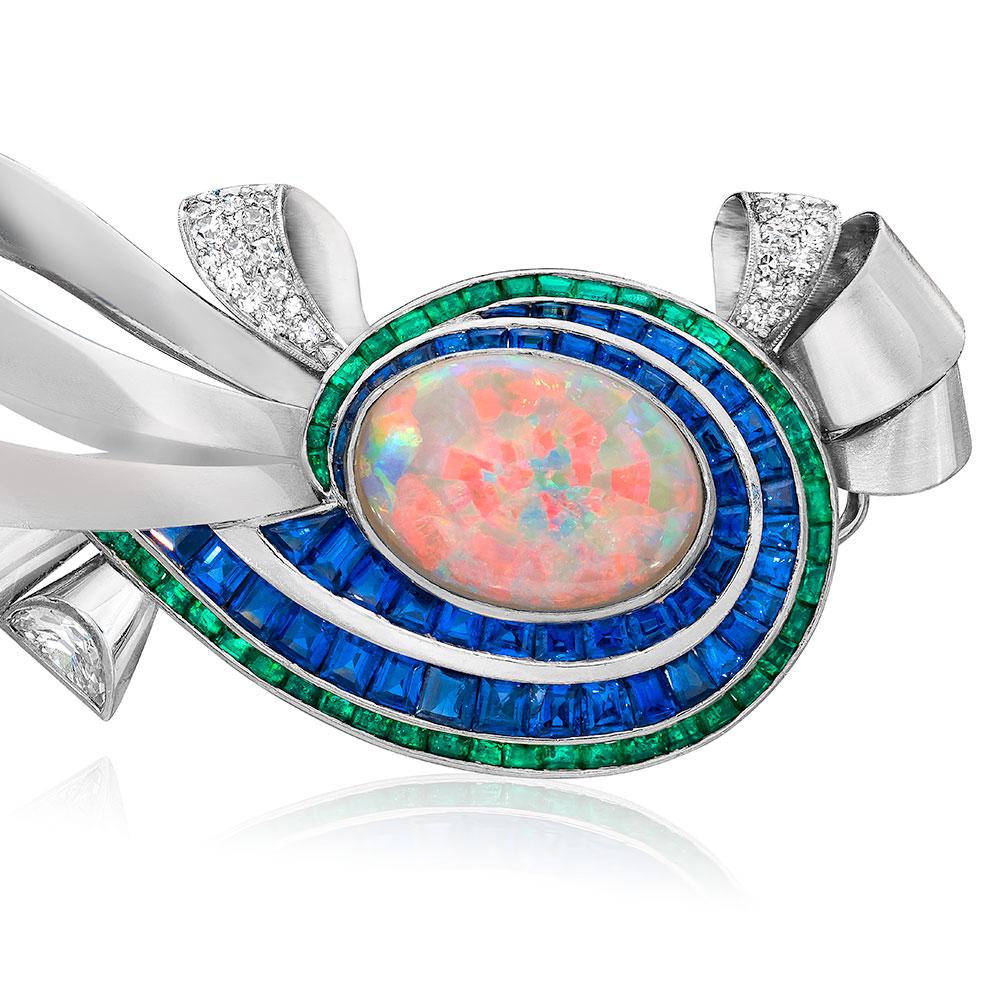 A swirling array of gemstones are set in this elegant mid century brooch. The center features a 5.73 carat oval Opal. Two rows of graduating calibre cut Blue Sapphires weighing 6.75 carats, and one row of calibre cut Emeralds weighing 2.31 carats 