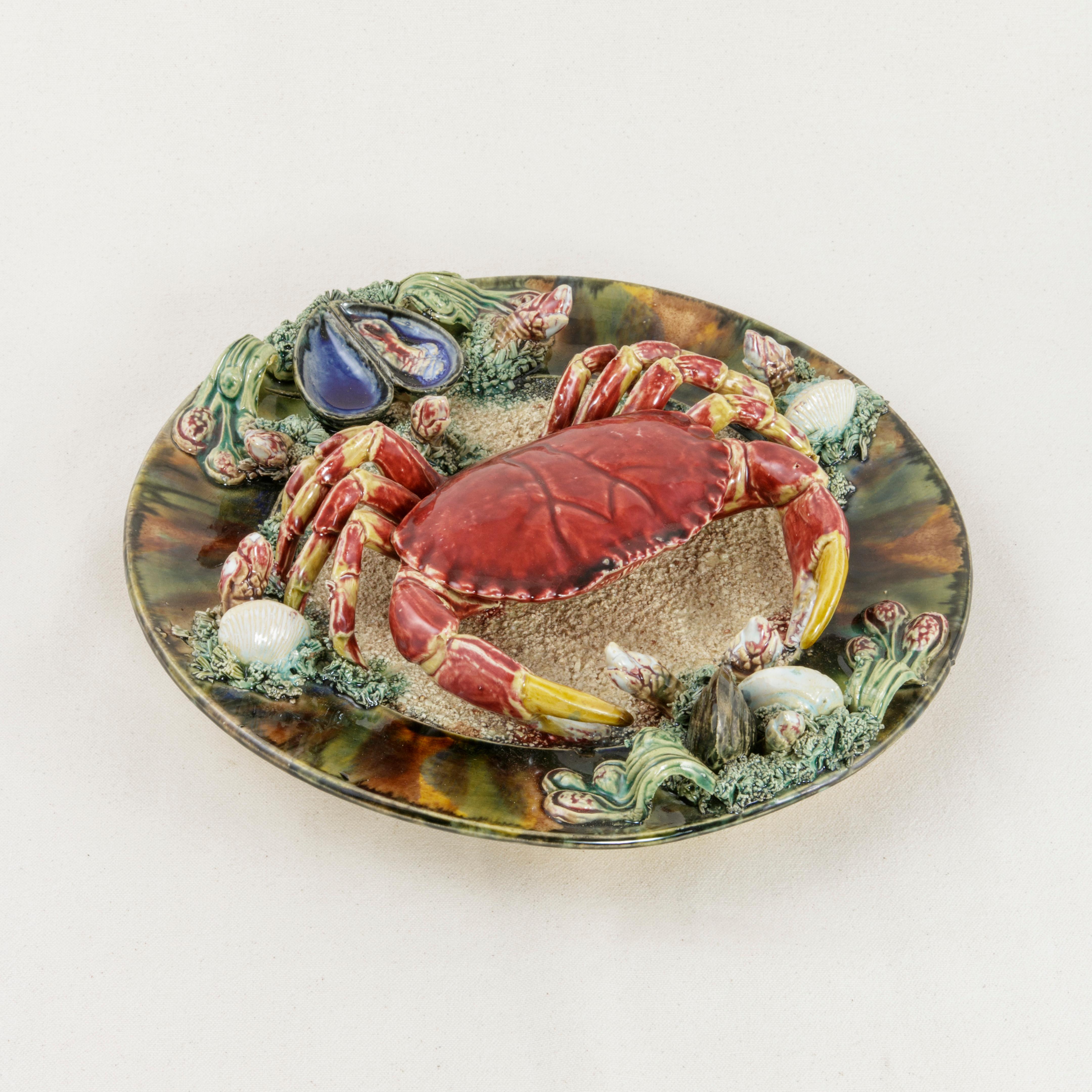 This large Portuguese Majolica Palissy style plate from the mid-20th century is glazed with vivid colors and features a hyper-realistic high-relief crab occupying the center of the plate. The crab is surrounded by elements of the sea such as mussels