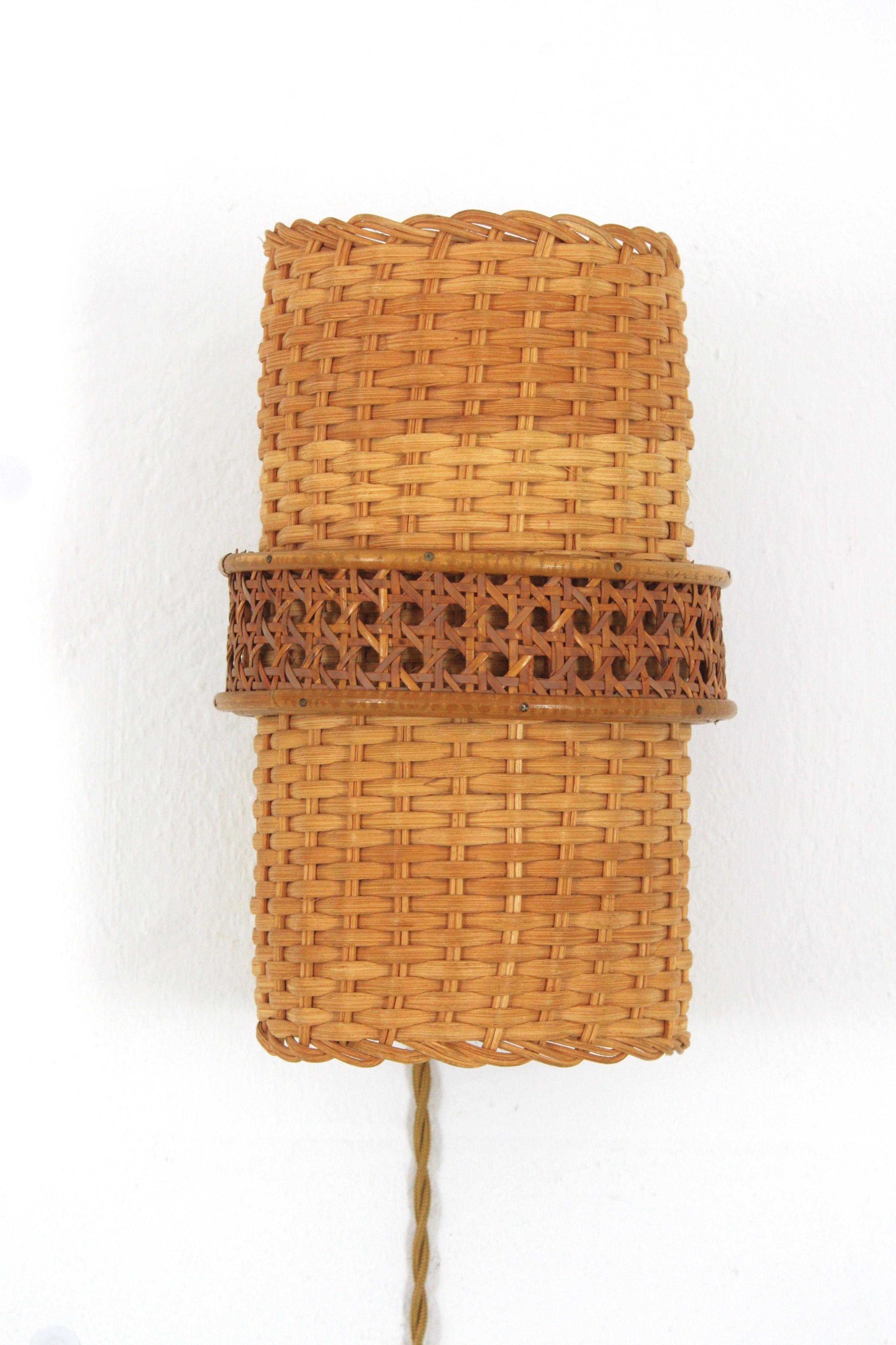 Moderner Wandleuchter aus Rattan und geflochtener Weide aus der Jahrhundertmitte. Spanien, 1950er Jahre.
Diese Wandleuchte aus handgeflochtenem Weidengeflecht hat ein auffälliges zylindrisches Design, das von einem Rattanring mit Canage-Detail als