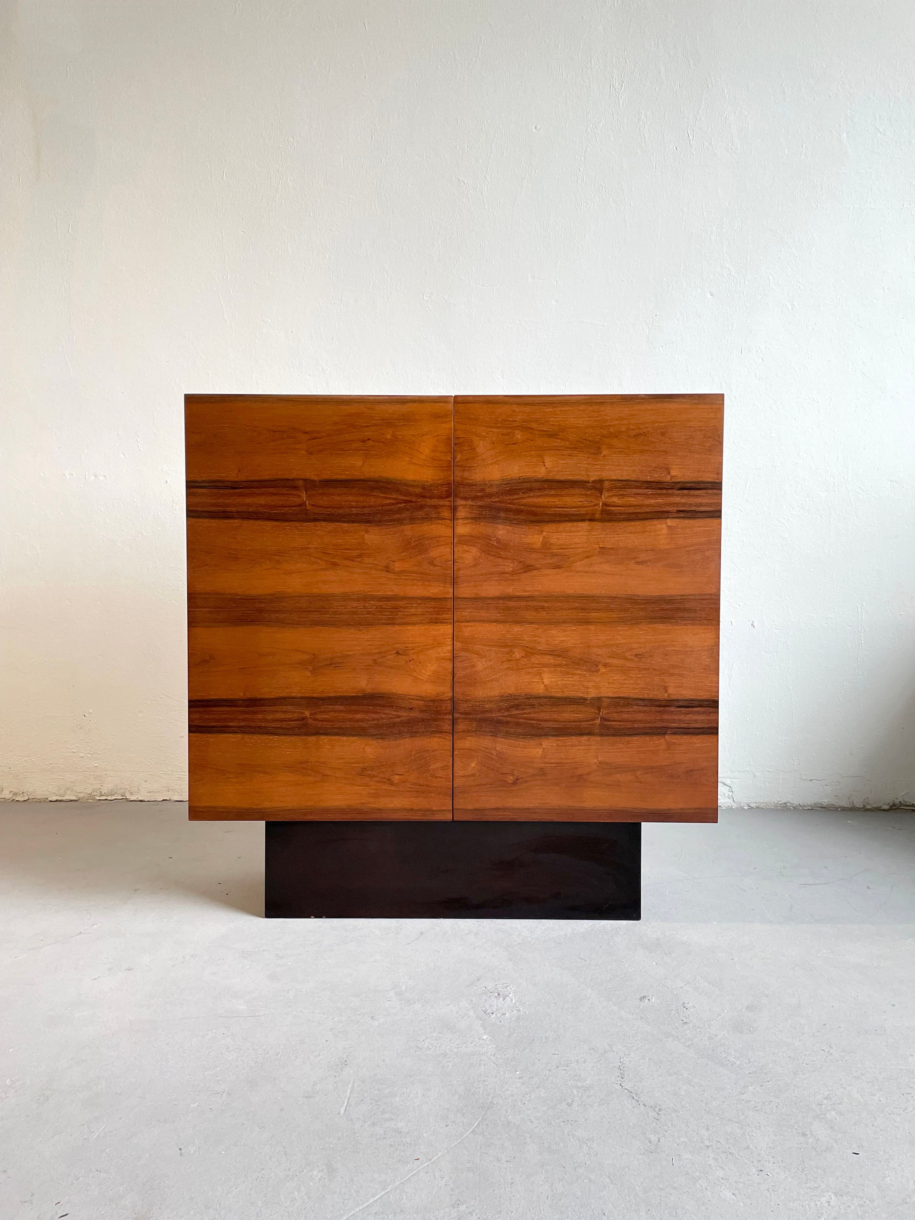 This high-end midcentury cabinet, made of rosewood, is manufactured in Germany in the 1970s.

Very sophisticated timeless Minimalist design that features front door made of rosewood and the glossy black wooden base and frame.
Storage space is
