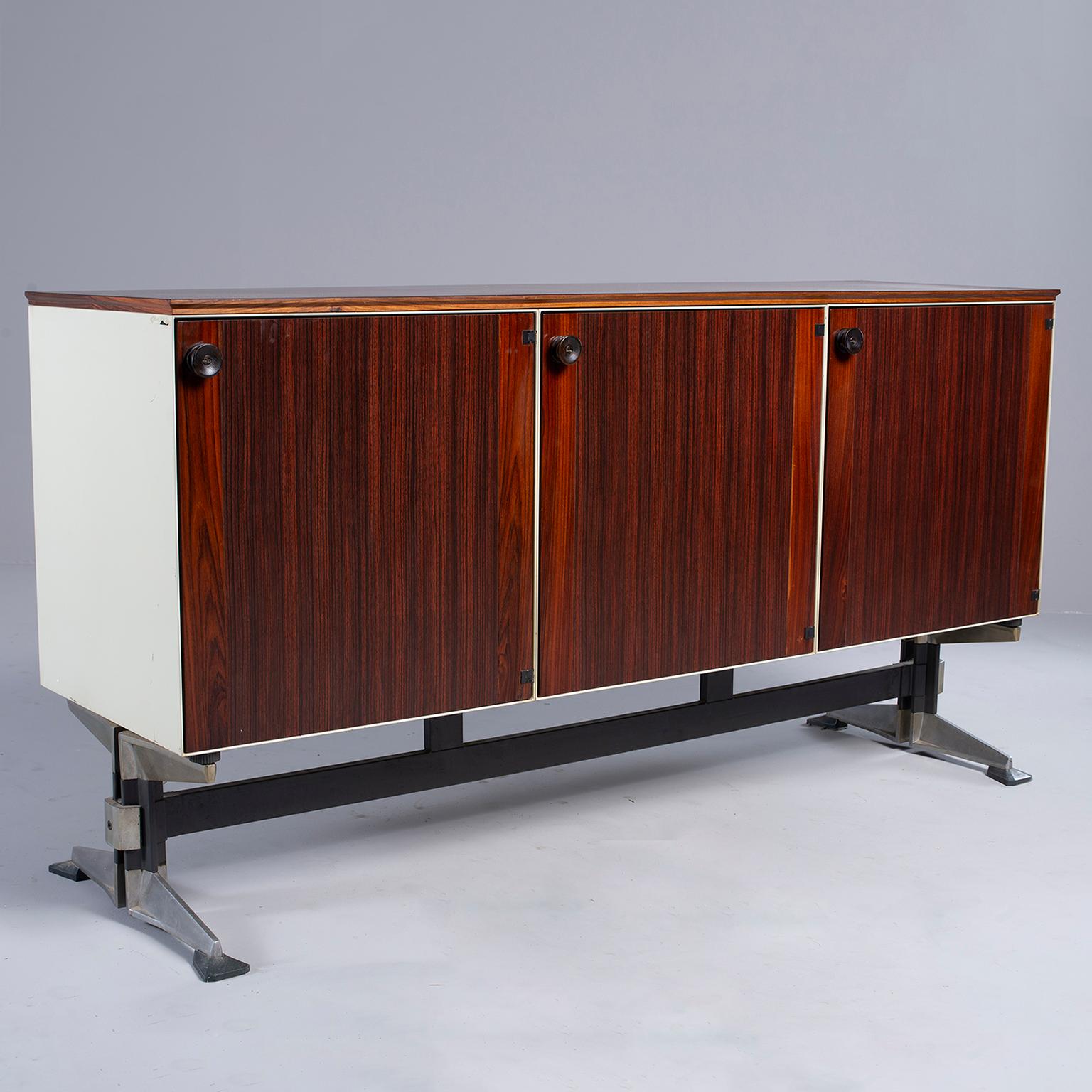 Cabinet or credenza has rosewood veneer front and top with three hinged door compartments with internal shelves, circa 1960s. Black stretcher with splayed metal feet. Sides and back of cabinet have a painted ivory colored finish. Depth measurement