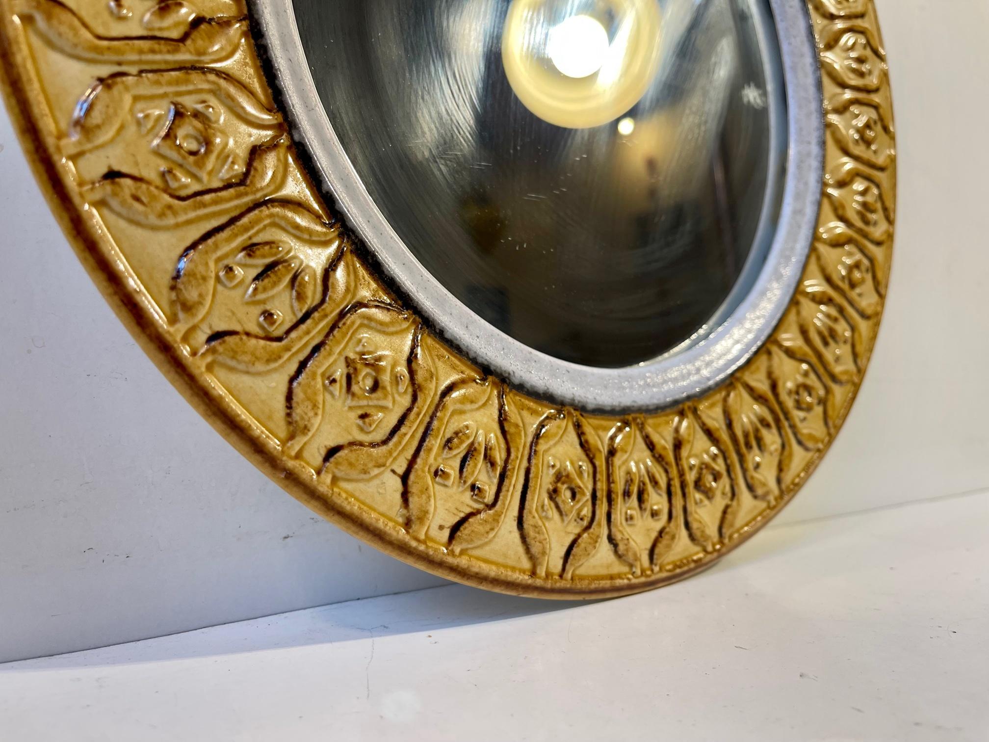 A decorative circular wall mirror in ocre yellow glazed ceramic. It was made in Italy during the 1970s, probably by Bitossi. It has no markings except from a partial price tag from the Danish retailer 'Magasin' that sold it. Measurements: Diameter:
