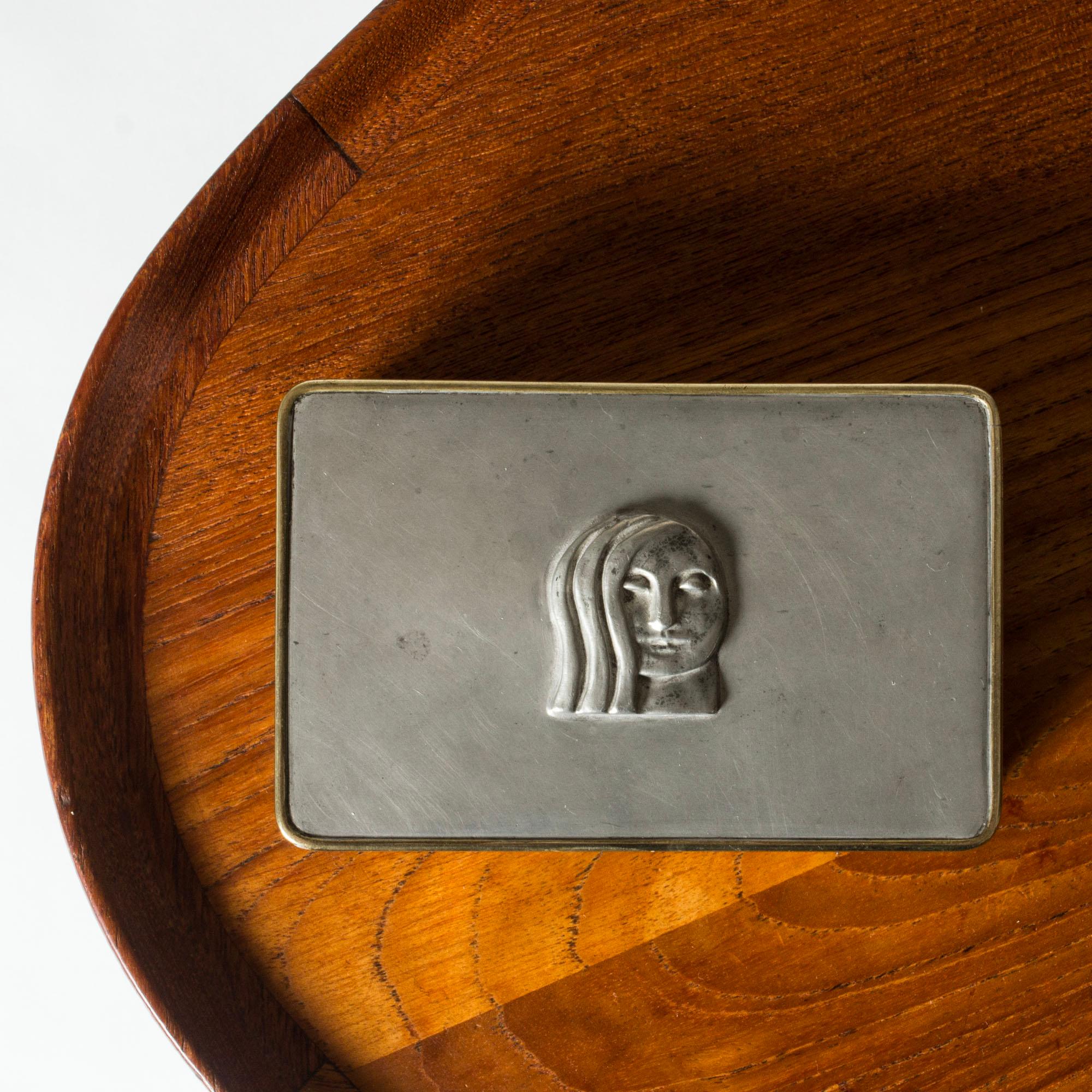 Elegant pewter case by Anna Petrus, with a line of brass around the lid. Decorated with a woman’s face in relief.
