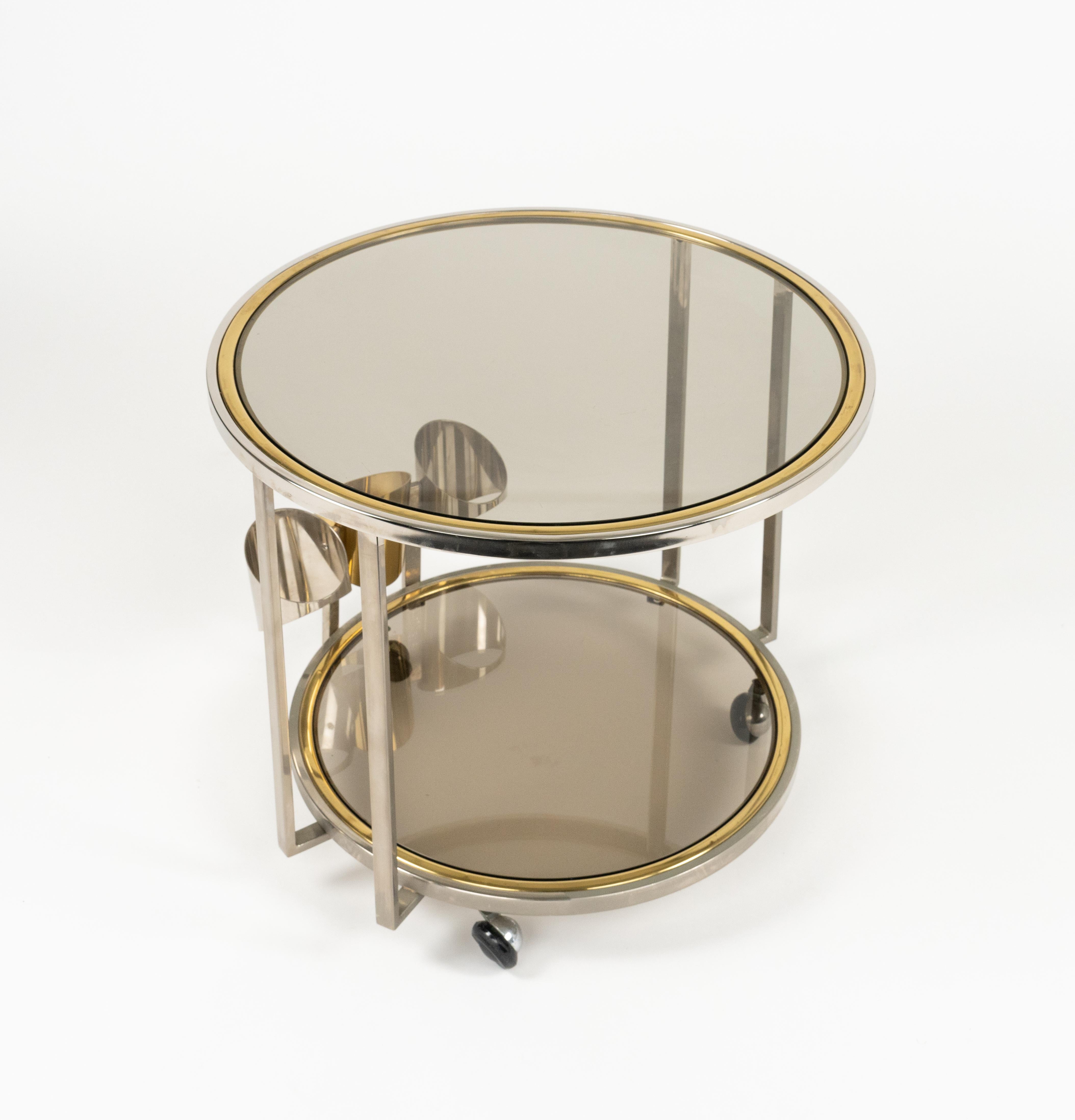 Etonnant chariot de bar rond à trois roues en laiton, chrome et verre fumé avec porte-bouteille dans le style de Romeo Rega.

Fabriqué en Italie dans les années 1970.

C'est un magnifique chariot de service qui épatera vos invités et qui est parfait