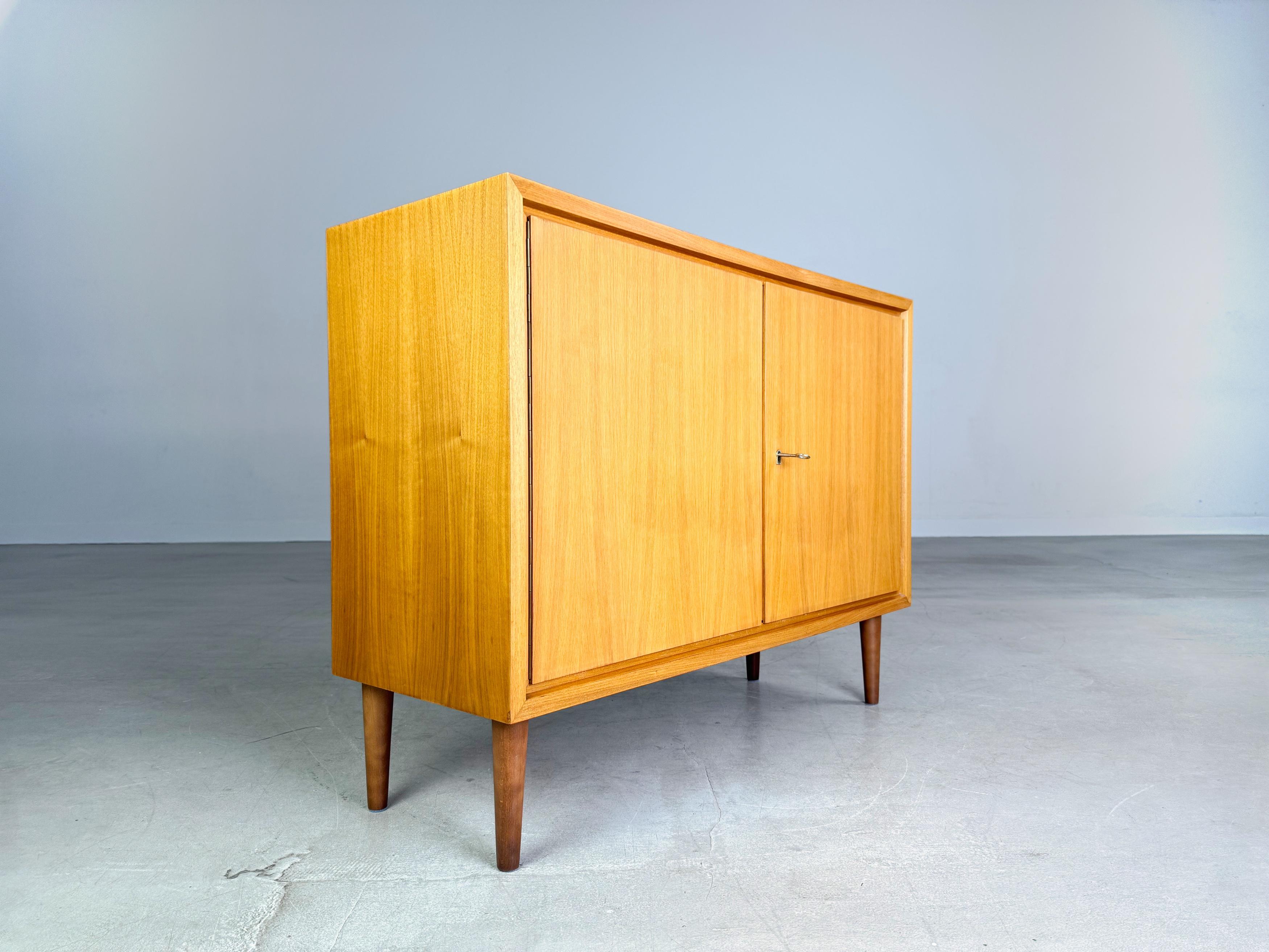 Wir bieten ein wunderschönes kleines WK Möbel Sideboard aus den 1960er Jahren zum Verkauf an. Das Sideboard verfügt über zwei Türen und wurde aus hochwertigem Nussbaumholz gefertigt. Die vier Füße sind aus gebeiztem Buchenholz und nachträglich