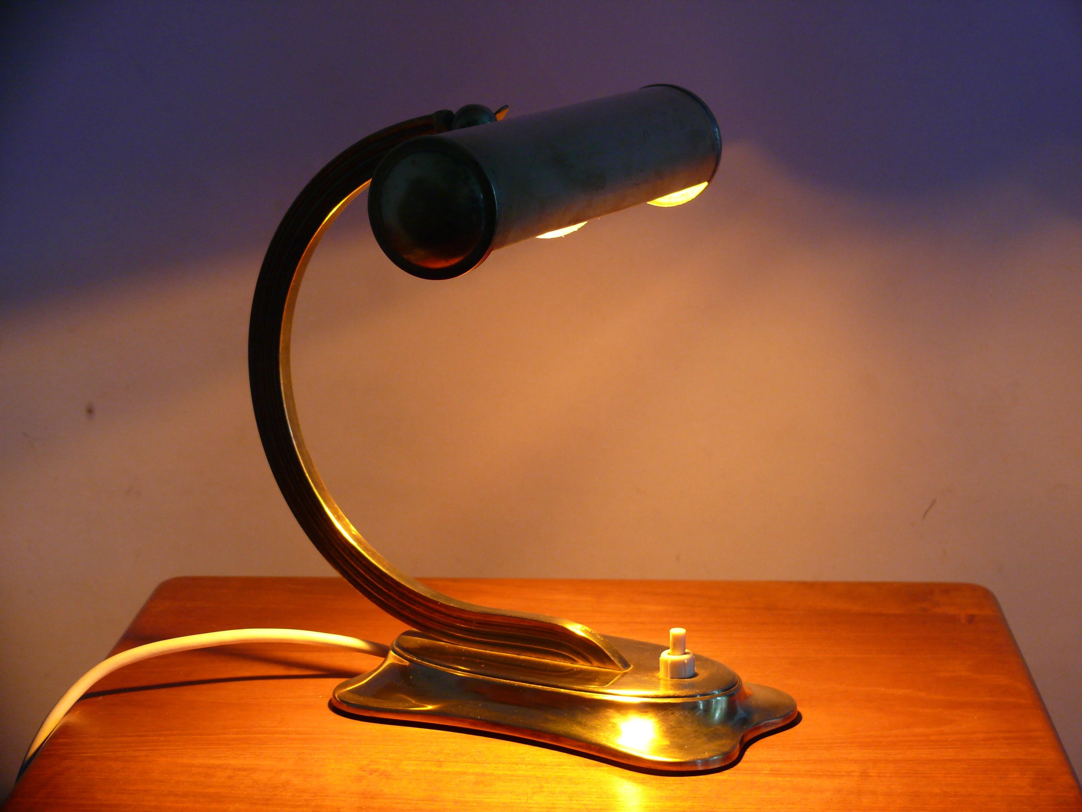 Small, charming brass table lamp from the 1950s. The base features an oval plate and a curved base. The curved arm features ribbed sides. The shade is adjustable using a thumbscrew, and a push-button switch is built into the base. The lamp is in