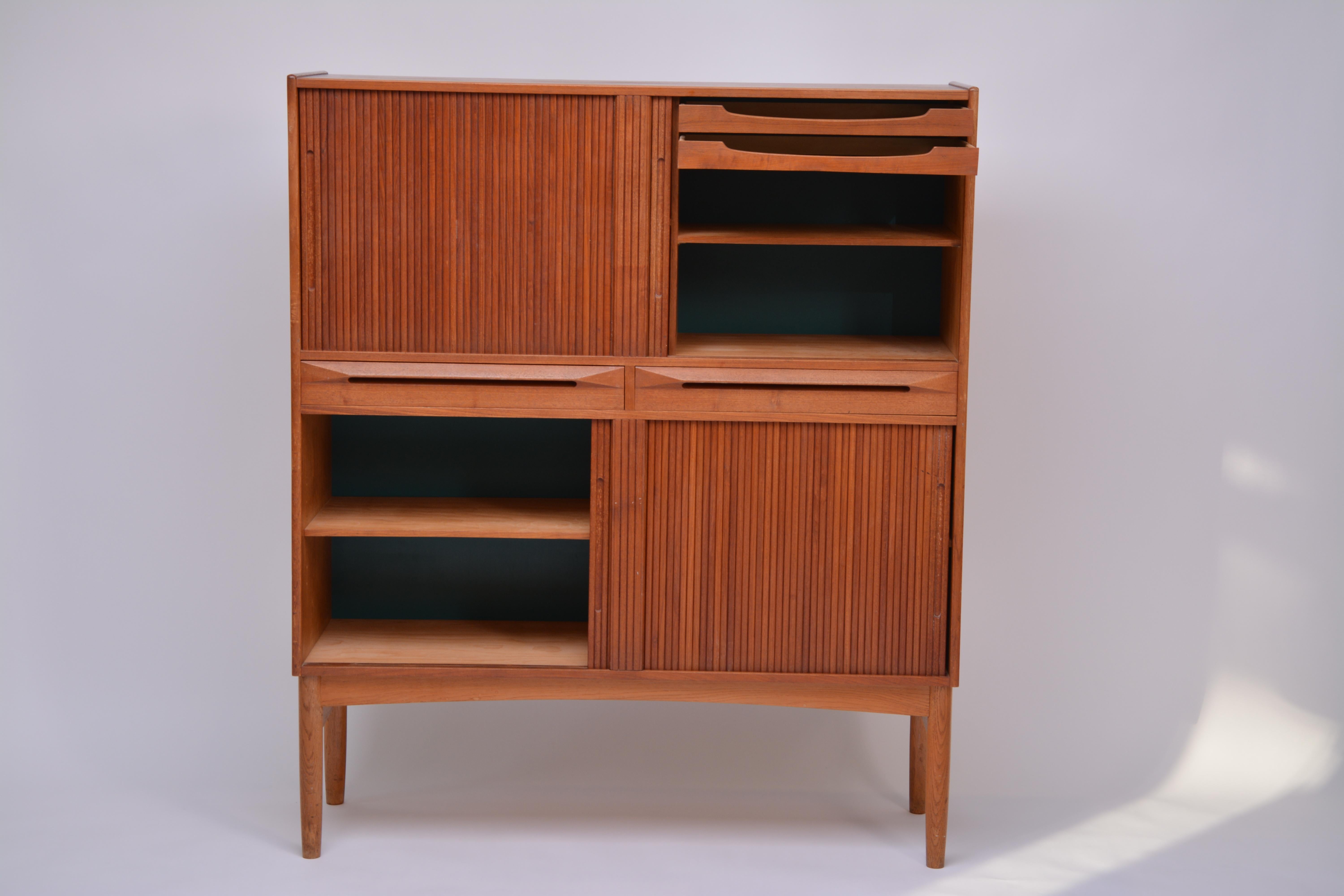 Tall teak cabinet with beech frame on beech shoes. Front with two drawers and four tambour doors. Inside with adjustable shelves and greenish blue lacquered back cover. Manufactured by Fredericia Stolefabrik.
