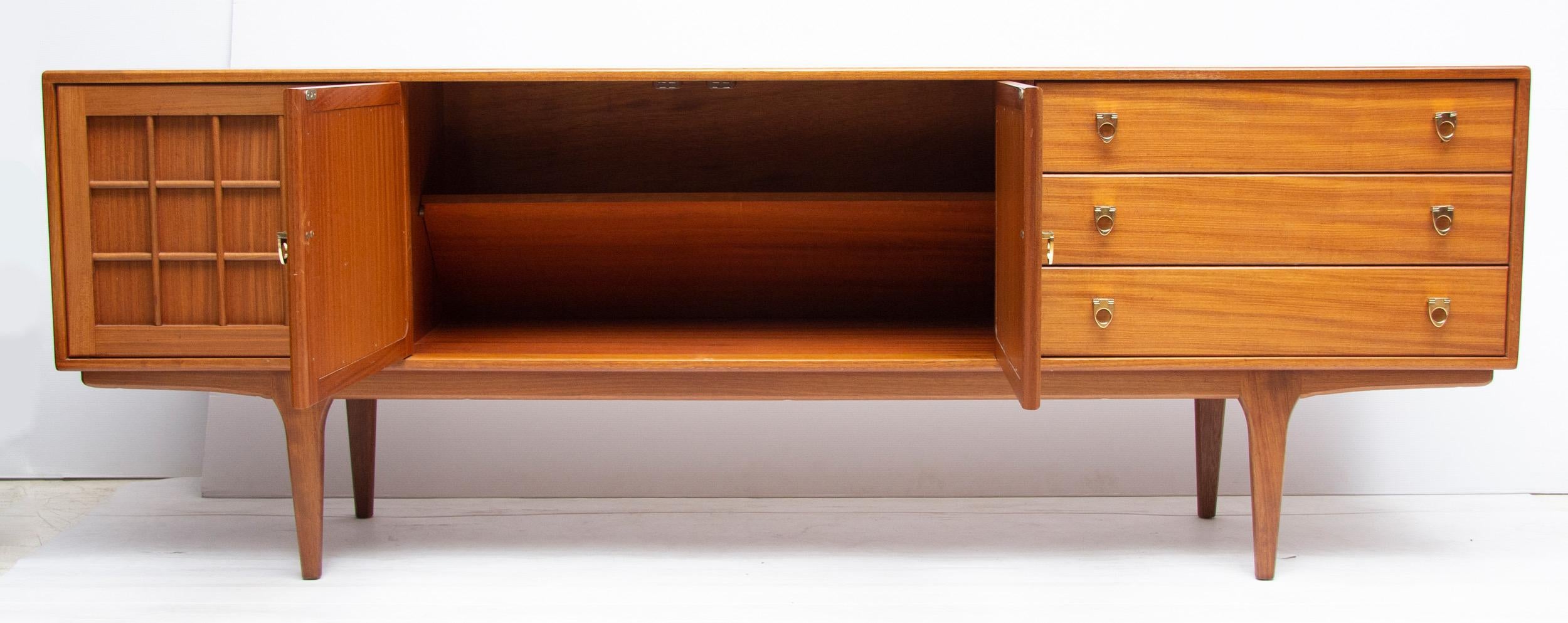 Midcentury sideboard credenza.
Beautiful golden teakwood sideboard.
Central, opposite opening panelled cupboard doors with cross bar decoration, flanked by another single cupboard and three drawers, with brass ring pull handles
Manufactured in