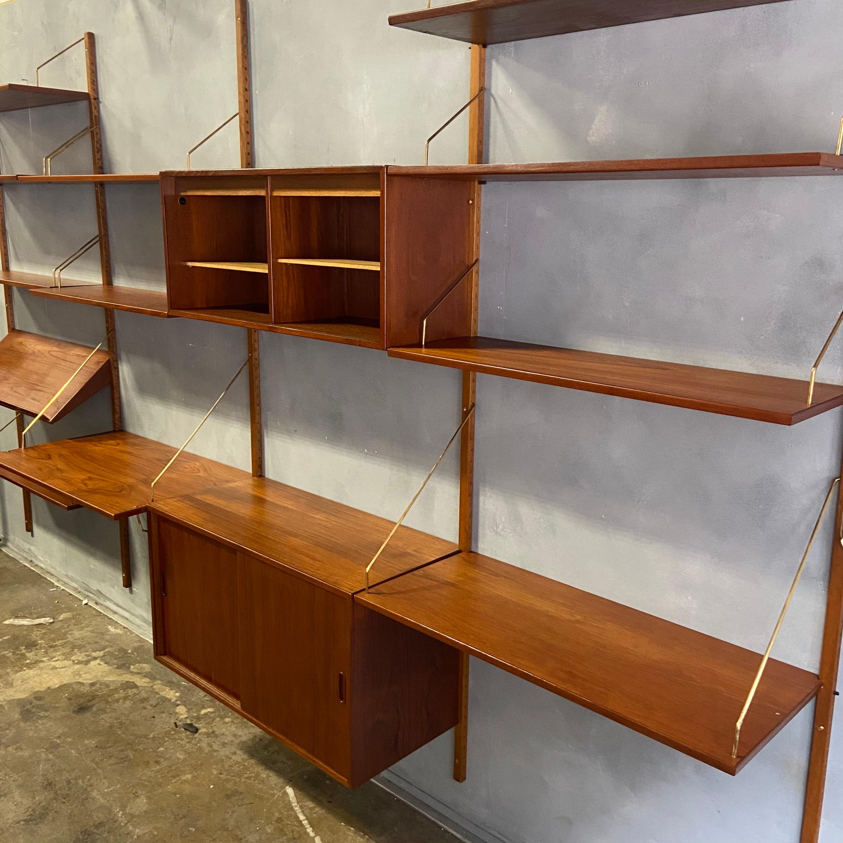Gorgeous teak wall unit with striking brass hanging elements. Wall unit has 4 bays that are 34.5 inches apart- rail to rail. Most shelves are 12'' deep and two are 9''. The wall unit features a cabinet with divided shelves, a desk that is 19.5''