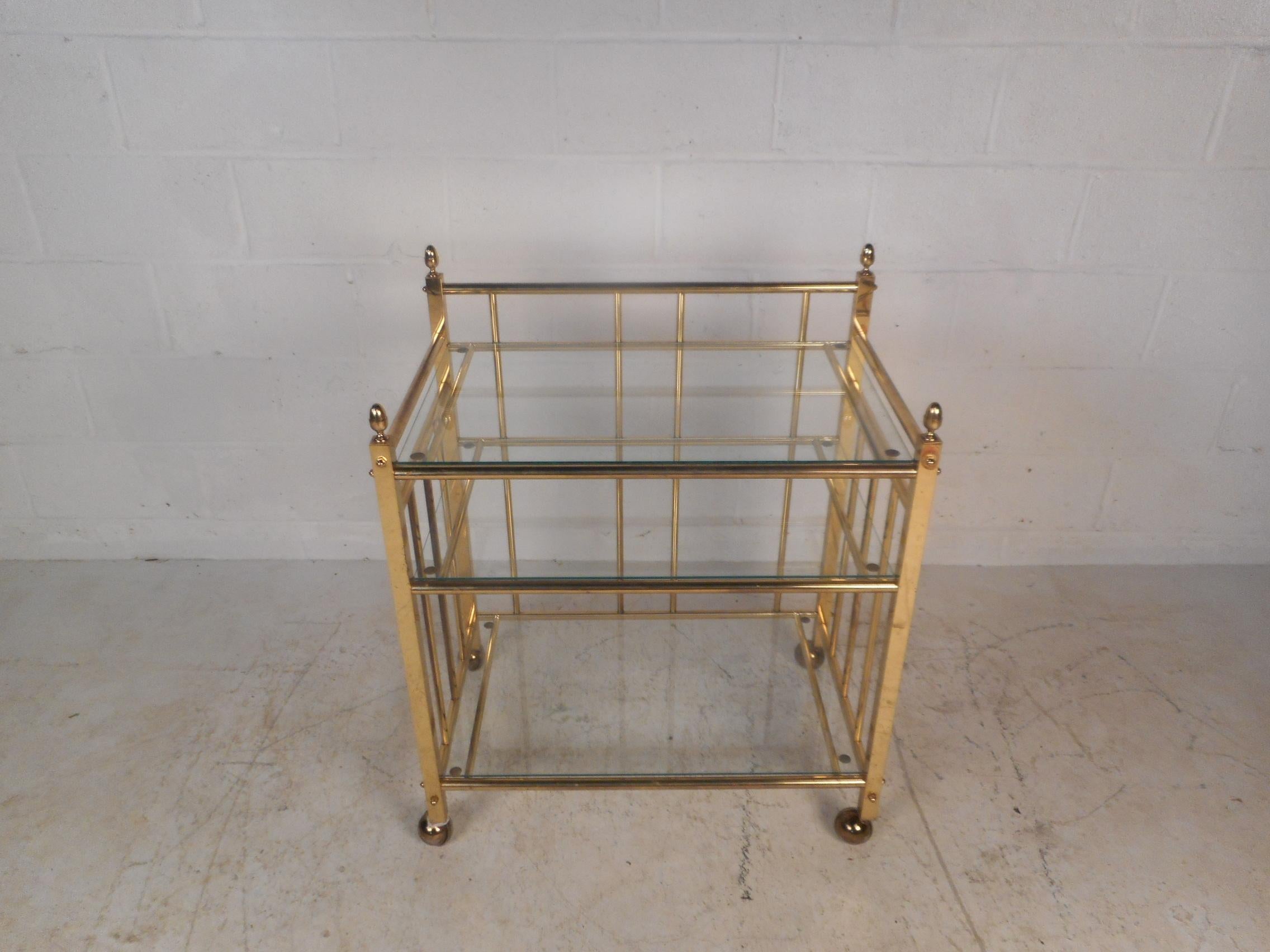 This sleek midcentury bar cart features a metal rod frame with three-tiers containing glass shelves, ensuring plenty of room for storage. This well designed serving cart has sculpted fixtures on top of each end and sits on top of four castors adding