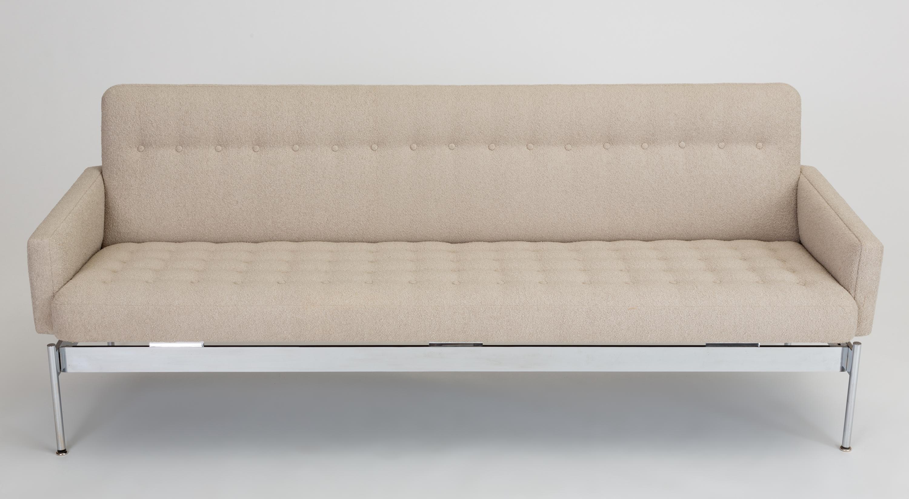 A Mid-Century Modern sofa with a slender profile sits atop a chrome base. The integrated cushion is button tufted, and a single row of buttons decorates the angled backrest. Each side is framed by a slim upholstered arm. The seat is angled to float